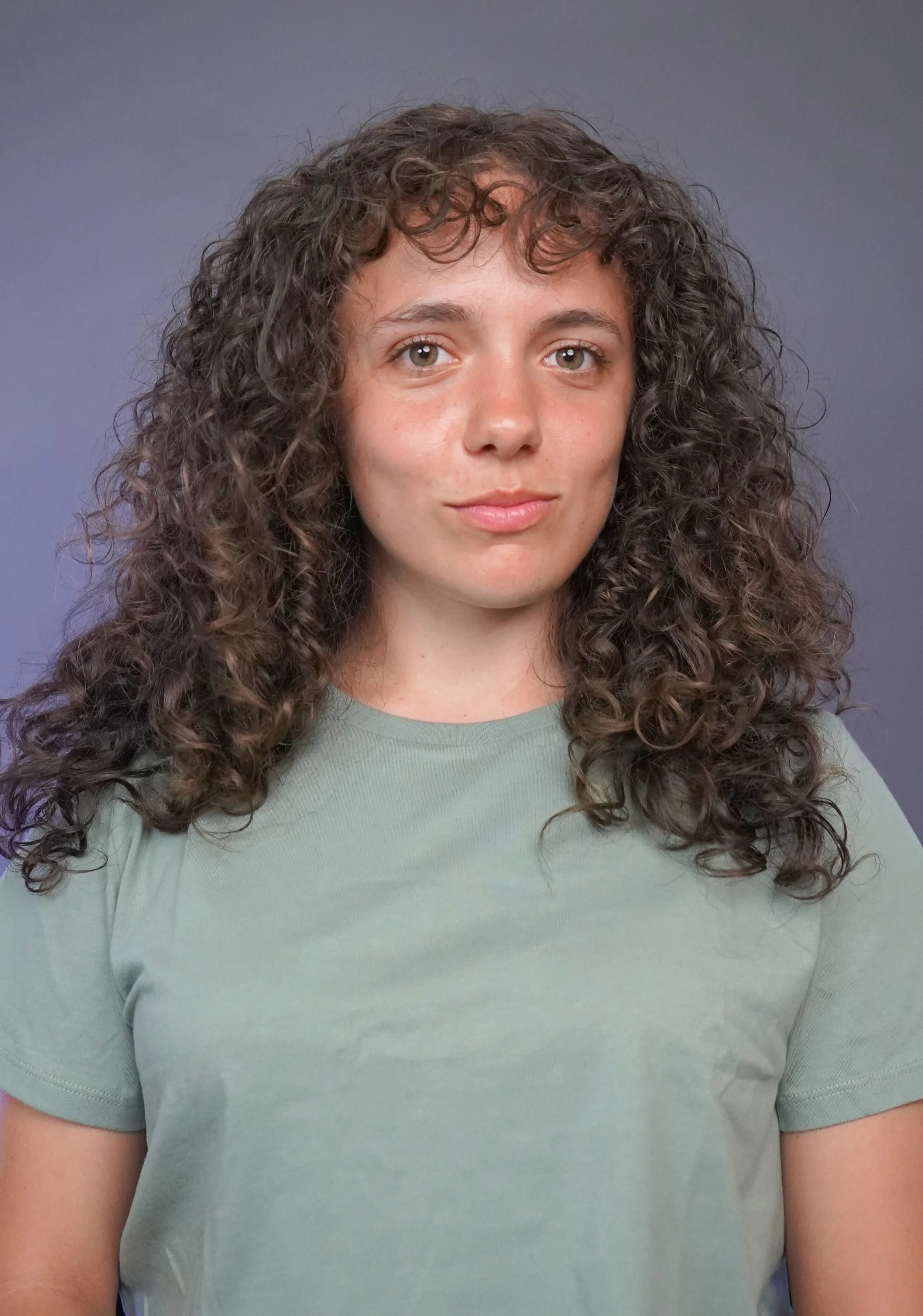 A young woman with long, curly brown hair and light skin wearing a light green T-shirt standing against a neutral background.