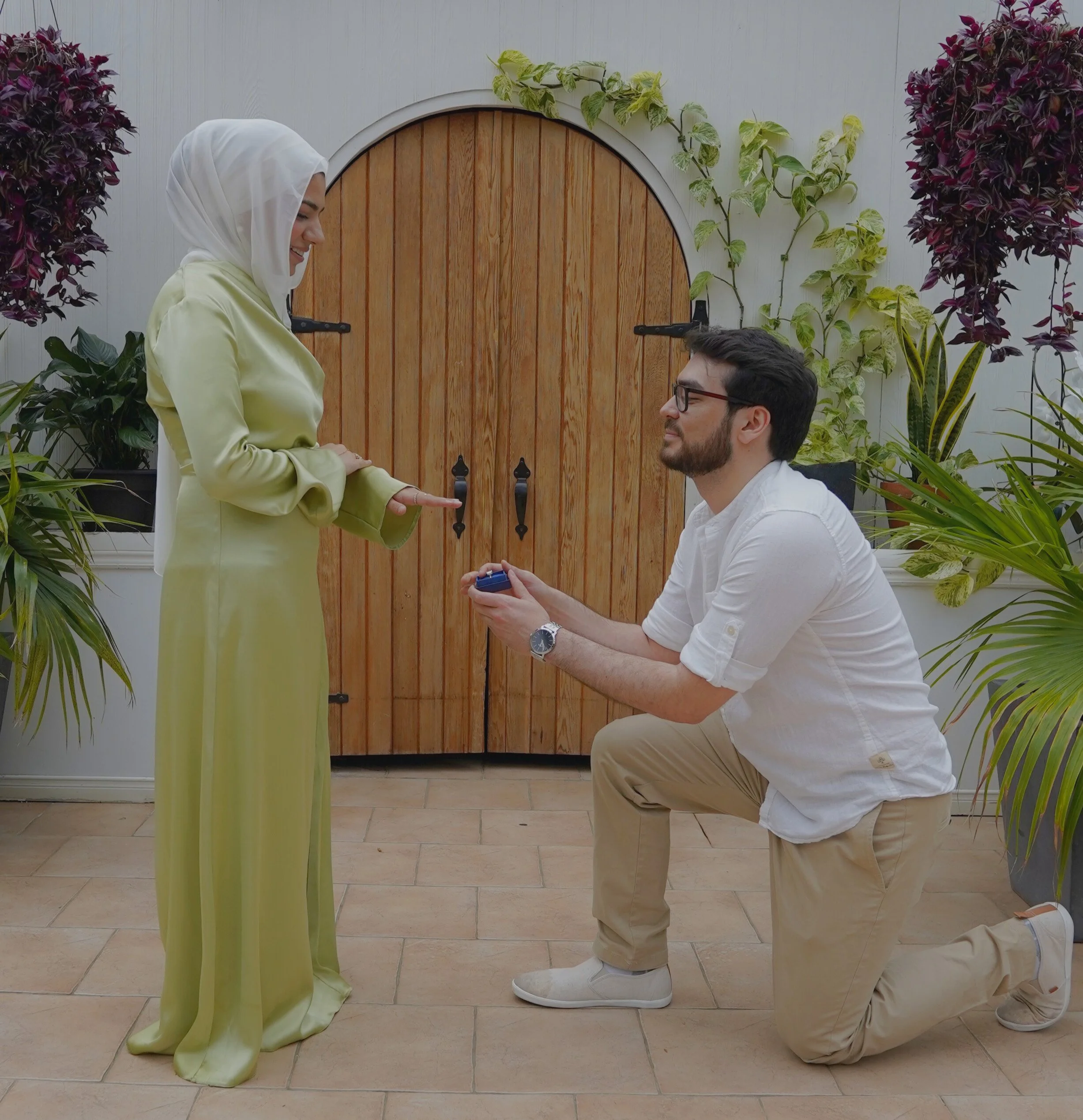 A man proposes to a woman with a ring, kneeling on one knee, in front of a garden door with surrounding plants.