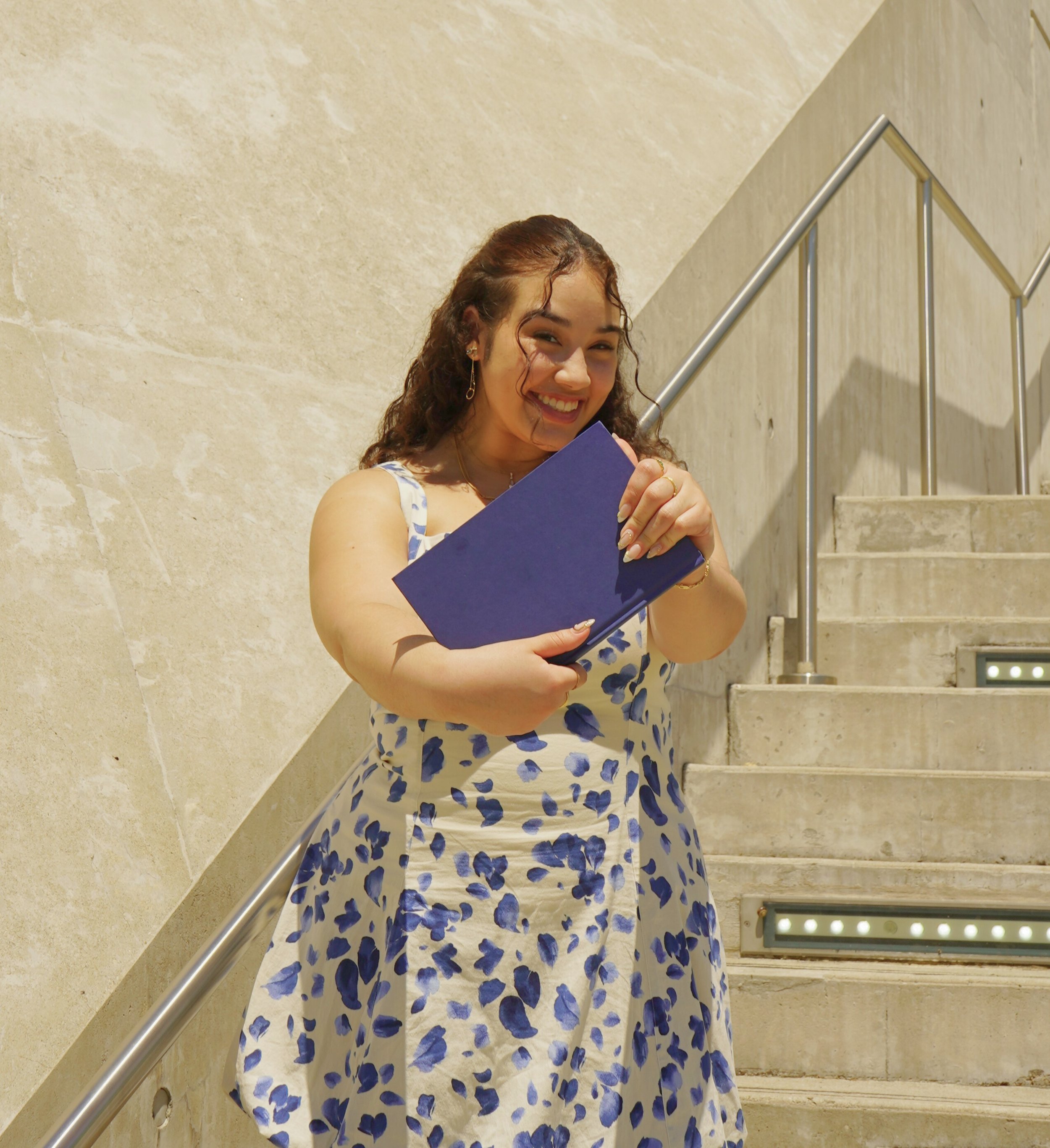 A young woman in a white dress with blue floral patterns, smiling and holding a blue folder, standing on a staircase with a beige stone wall and metal railing.