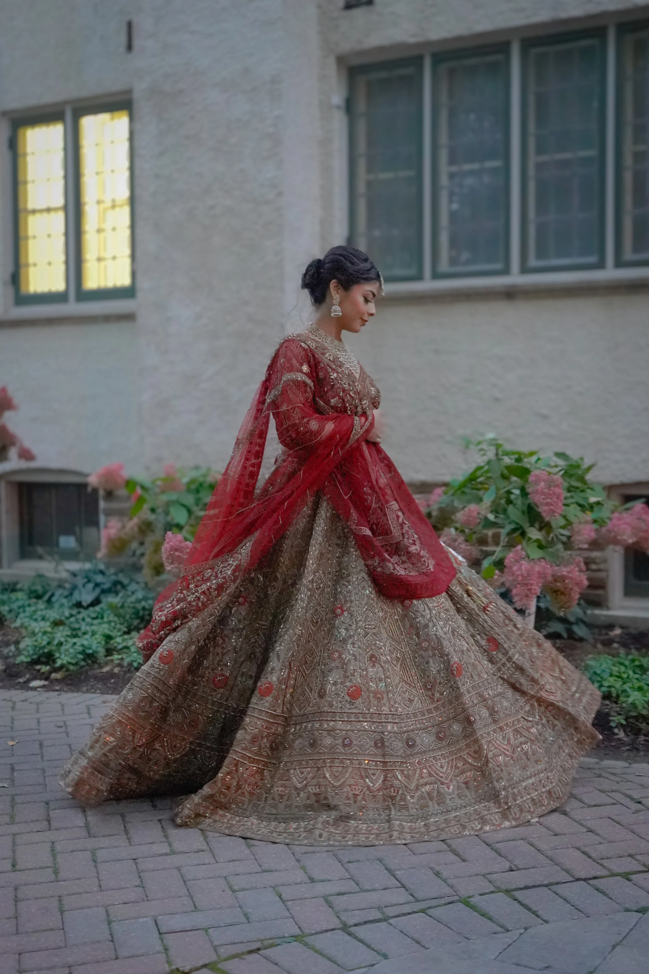 A woman wearing a traditional Indian bridal outfit in red and gold, posing outdoors near a garden with pink flowers.