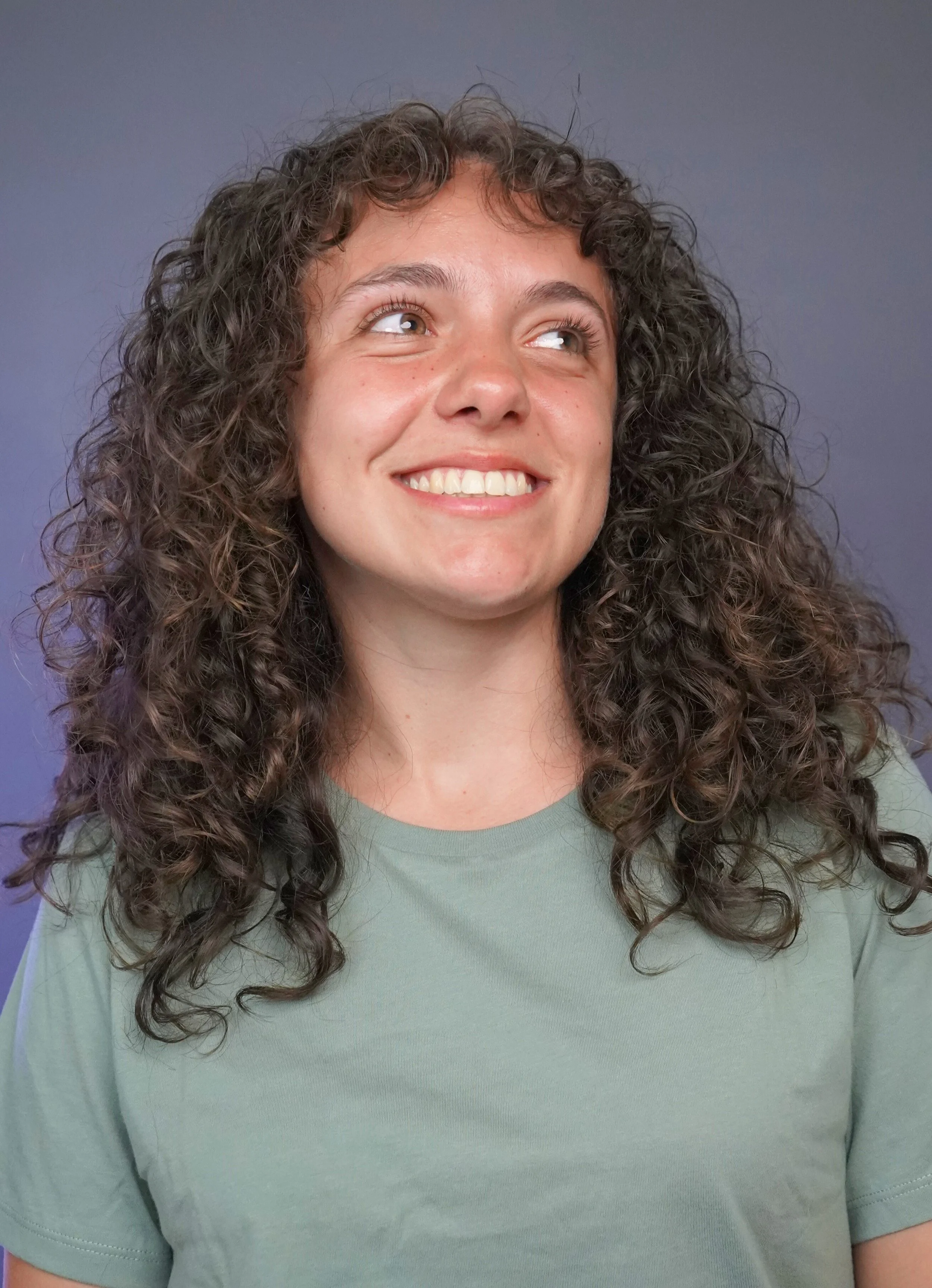A young woman with curly hair, smiling, wearing a green shirt, standing against a blue background.