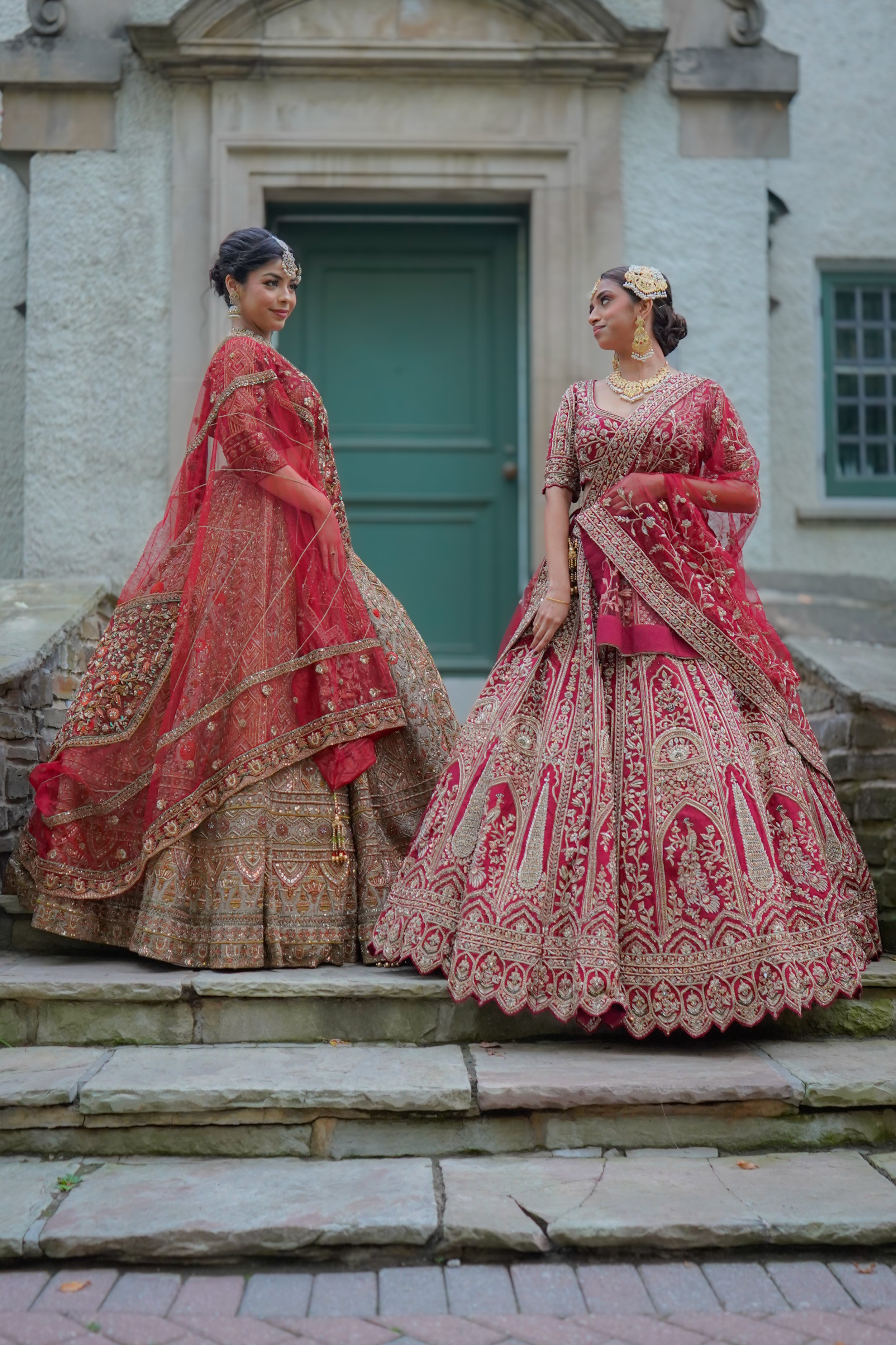 Two women wearing traditional Indian bridal sarees with intricate embroidery, standing on stone steps in front of a historic building with a green door.