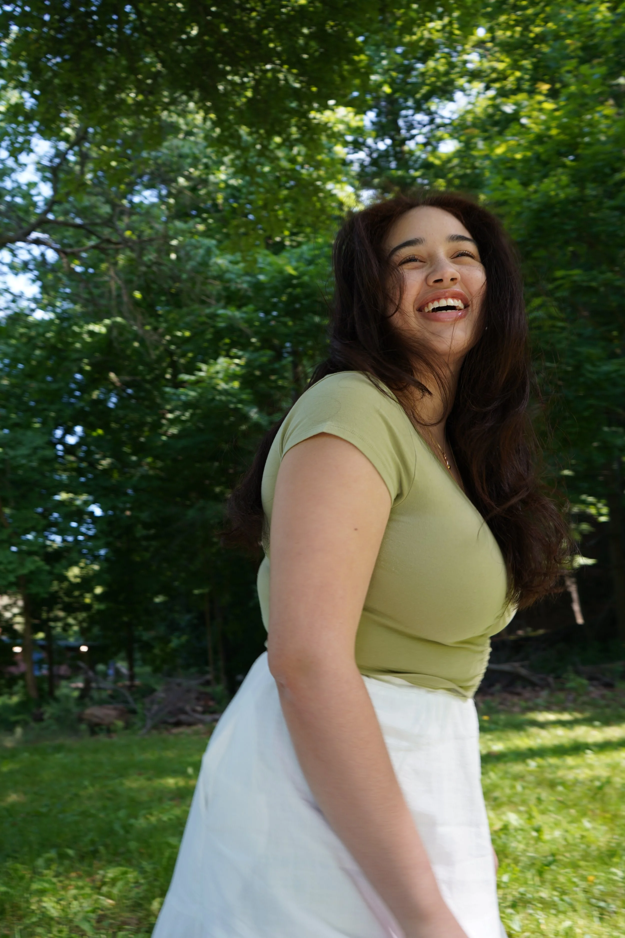 A woman with long dark hair, wearing a light green top and white skirt, laughing outdoors in a grassy area with trees in the background.