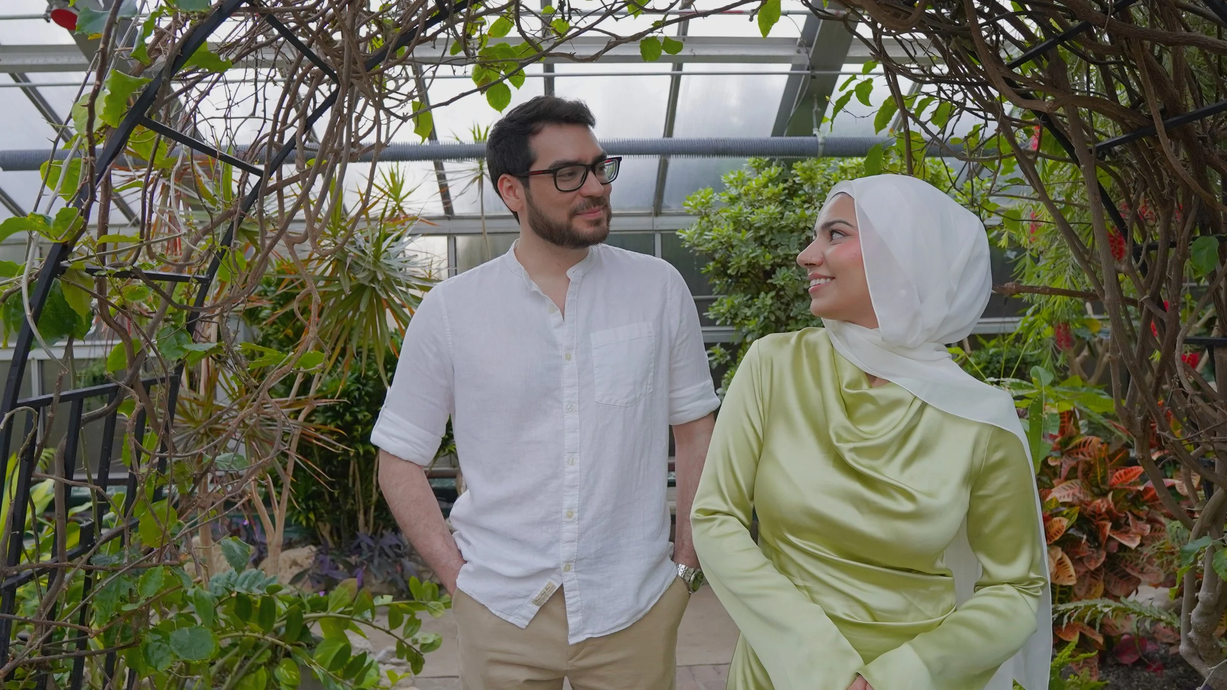 A man and woman are talking inside a greenhouse filled with plants. The man has dark hair, glasses, and a beard, and is wearing a white shirt. The woman is wearing a green dress and a white headscarf, and they are smiling at each other.