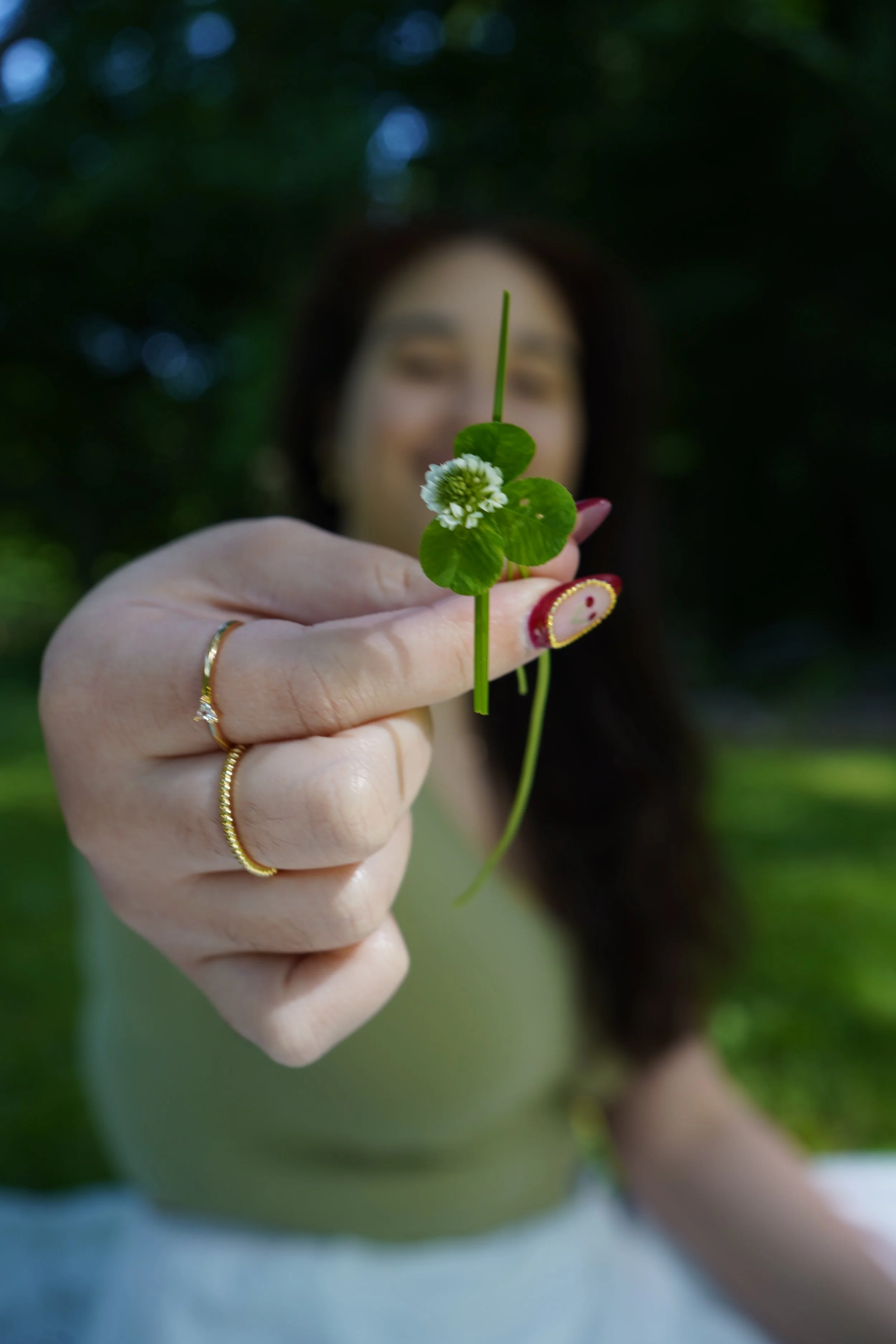A woman holding a small green plant with a white flower close to the camera, while sitting outdoors in a park or garden.