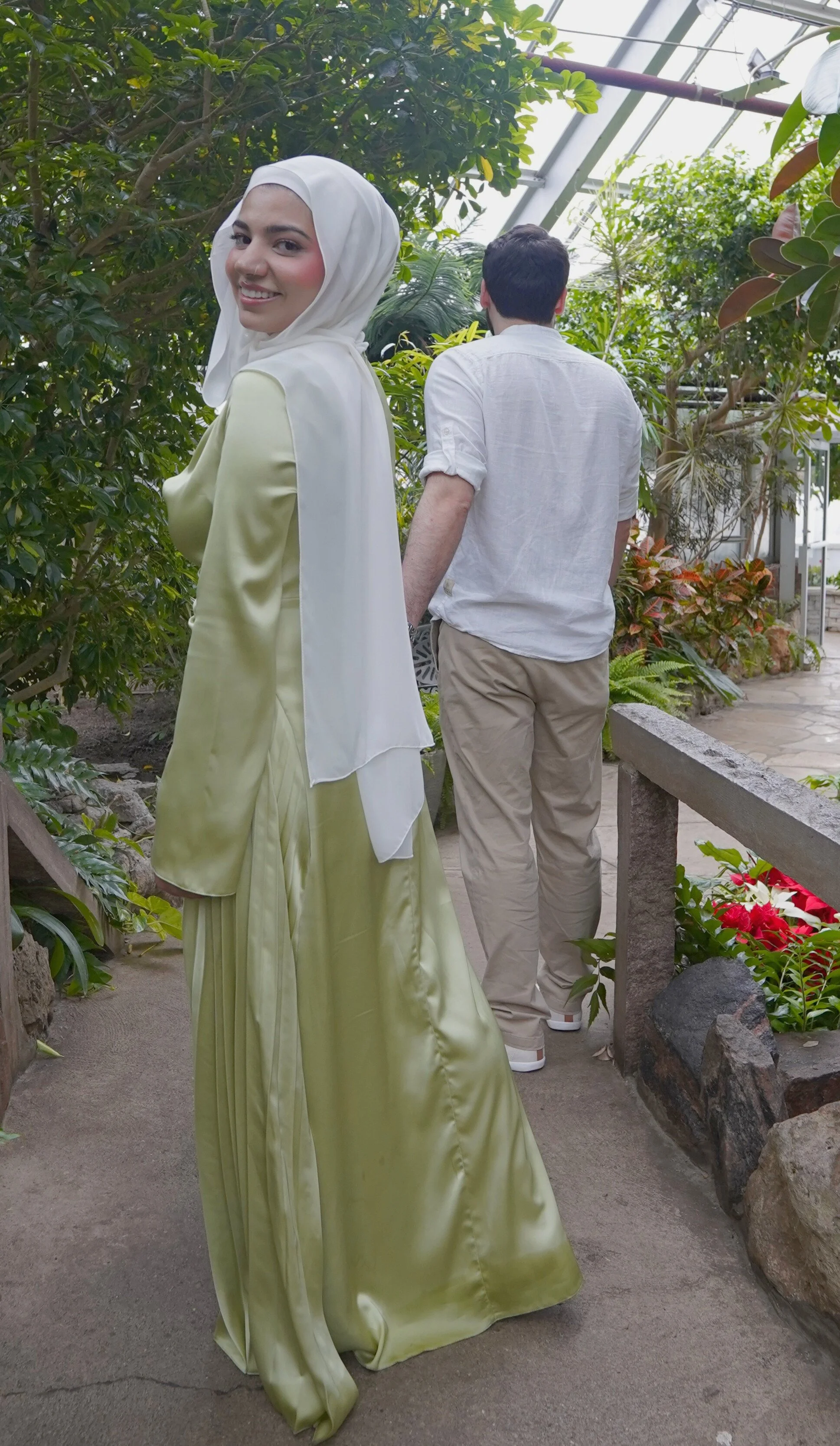 A woman wearing a white headscarf and a light green satin dress standing in a greenhouse with lush green plants and a man with dark hair in a white shirt and beige pants in the background.