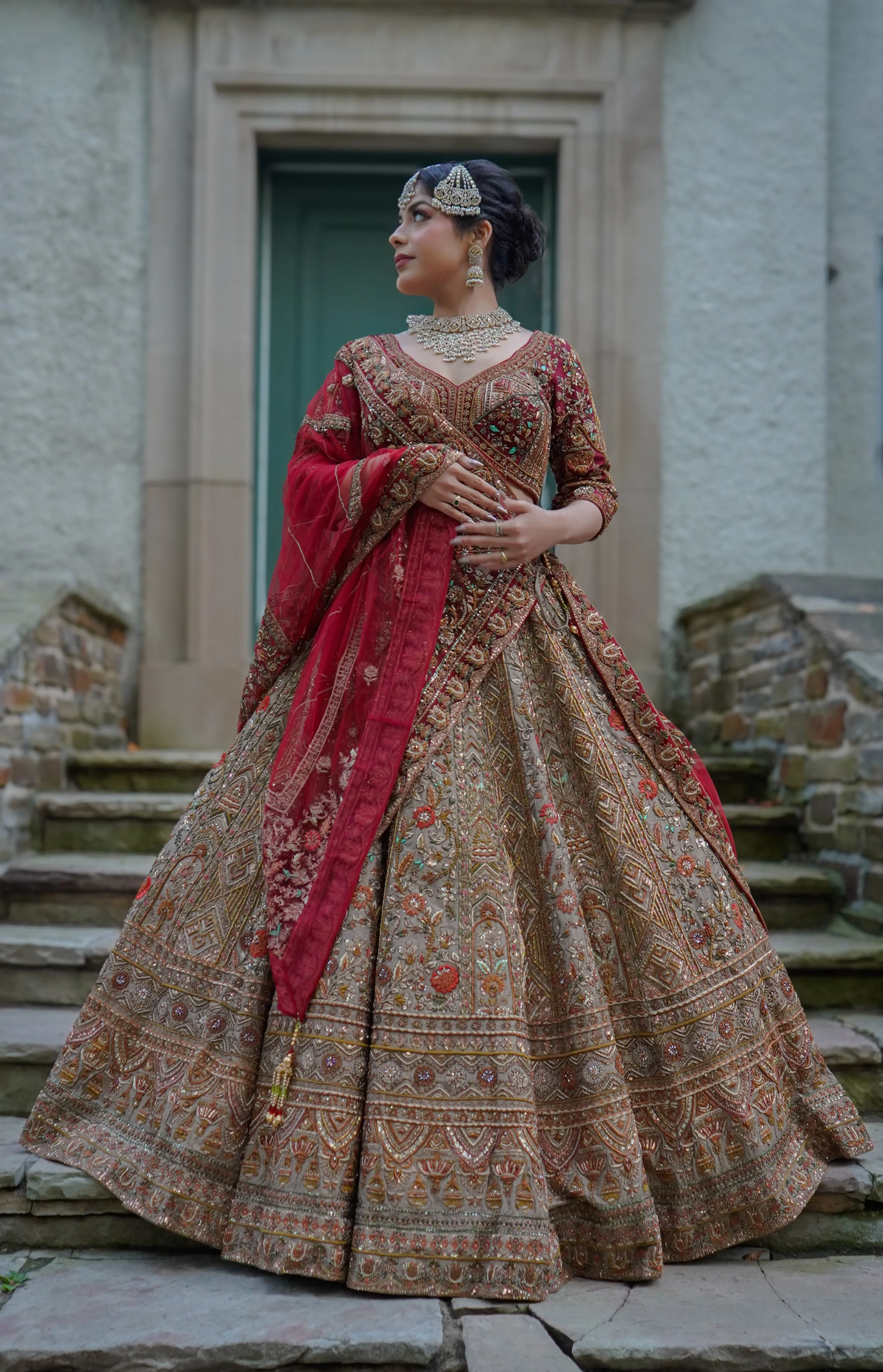 A woman dressed in an ornate red and gold traditional Indian bridal outfit, adorned with jewelry and standing on stone steps in front of a historic building.