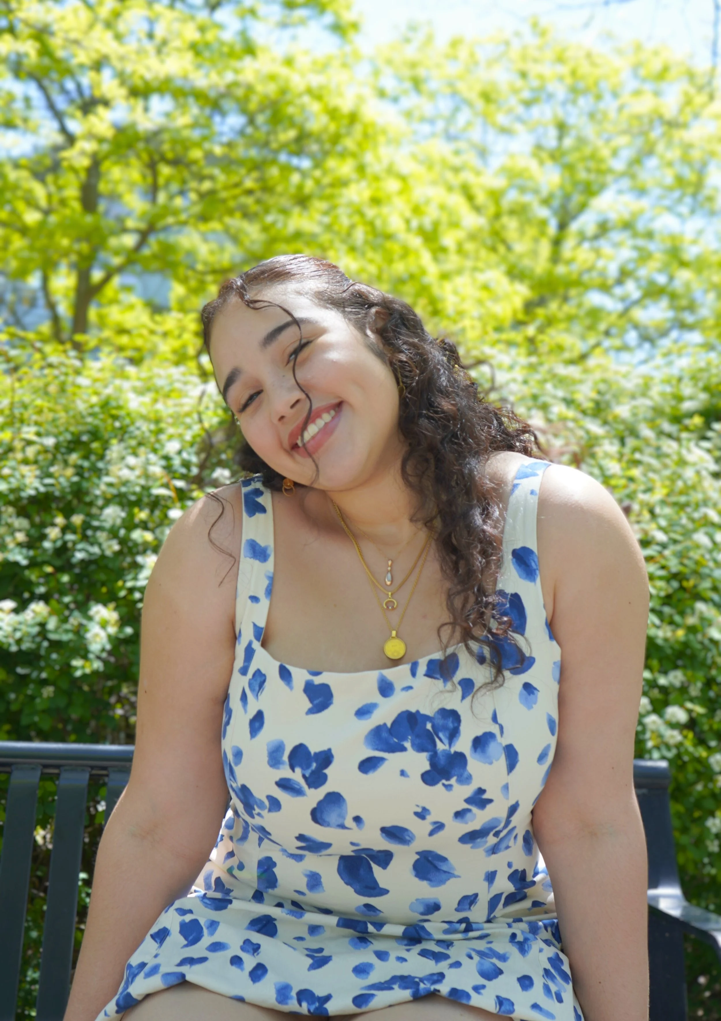 A young woman smiling outdoors on a sunny day, wearing a white dress with blue floral patterns, with lush green trees in the background.