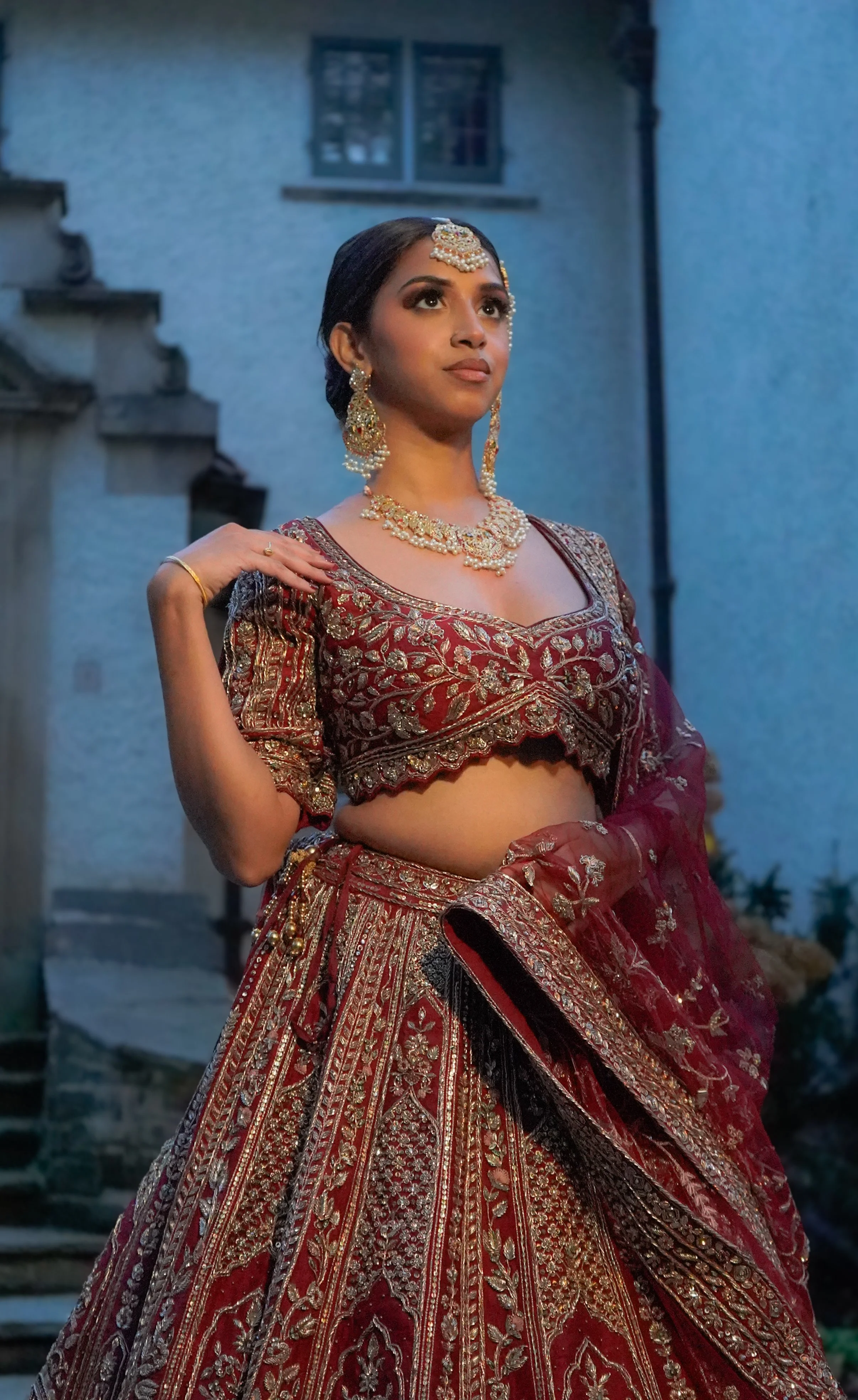 A woman in traditional Indian attire, wearing a richly embroidered maroon lehenga and choli with gold and silver embellishments, accessorized with jewelry including earrings, a necklace, and a headpiece. She stands outdoors in front of a building wit