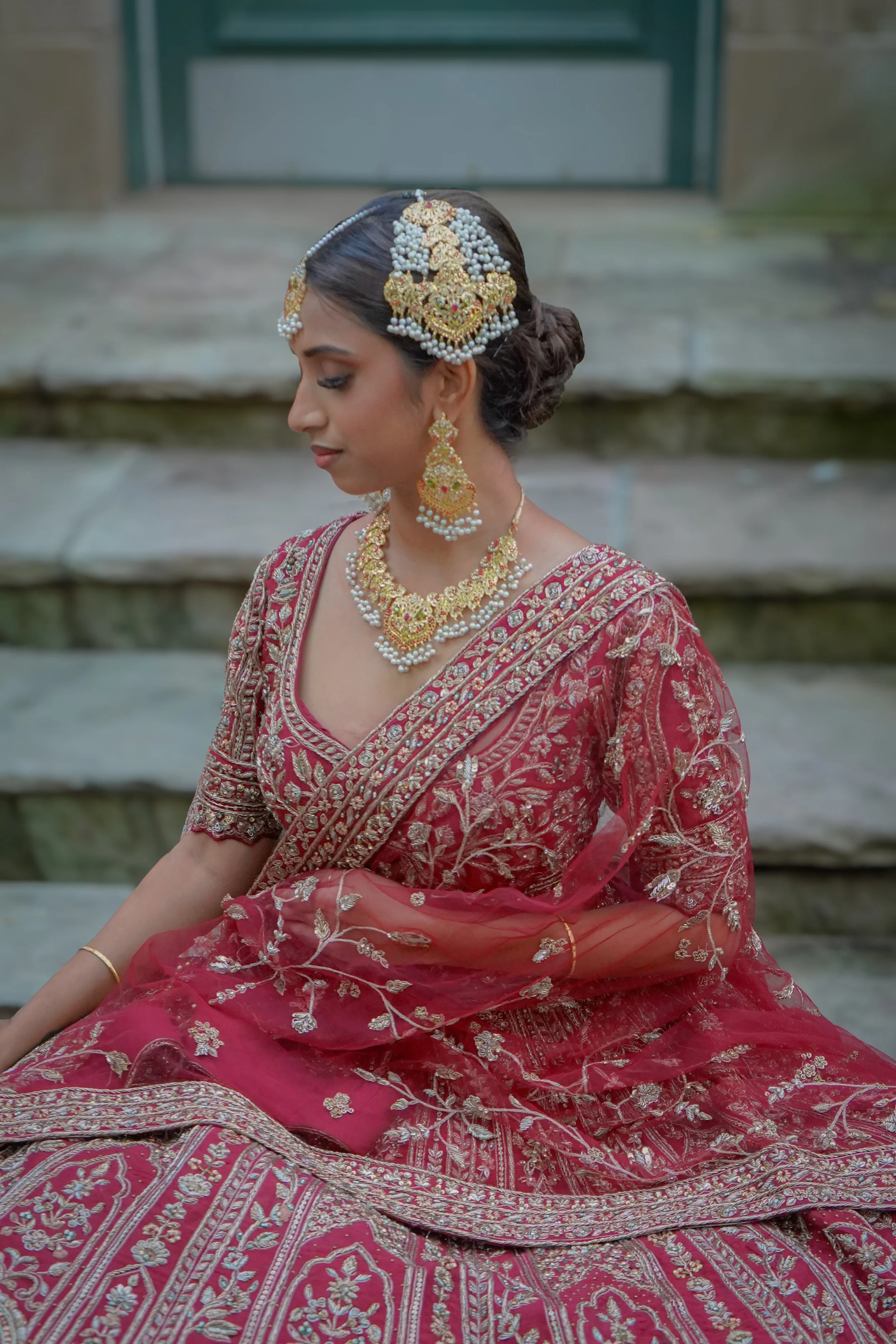 A woman dressed in traditional Indian bridal attire with intricate jewelry and a red embroidered saree, sitting on stone steps.