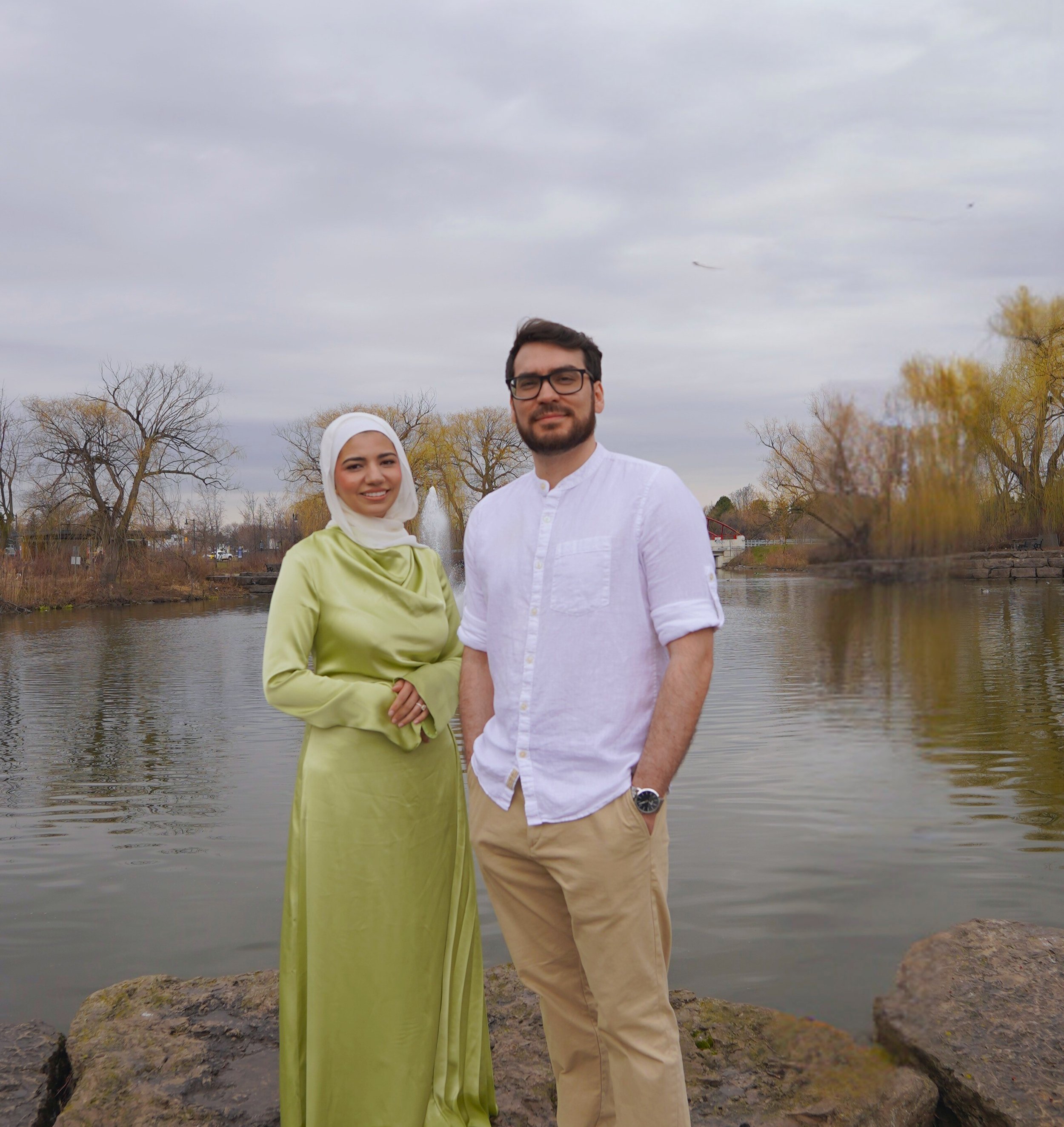 A man and woman standing by a pond with trees and a fountain in the background during a cloudy day.