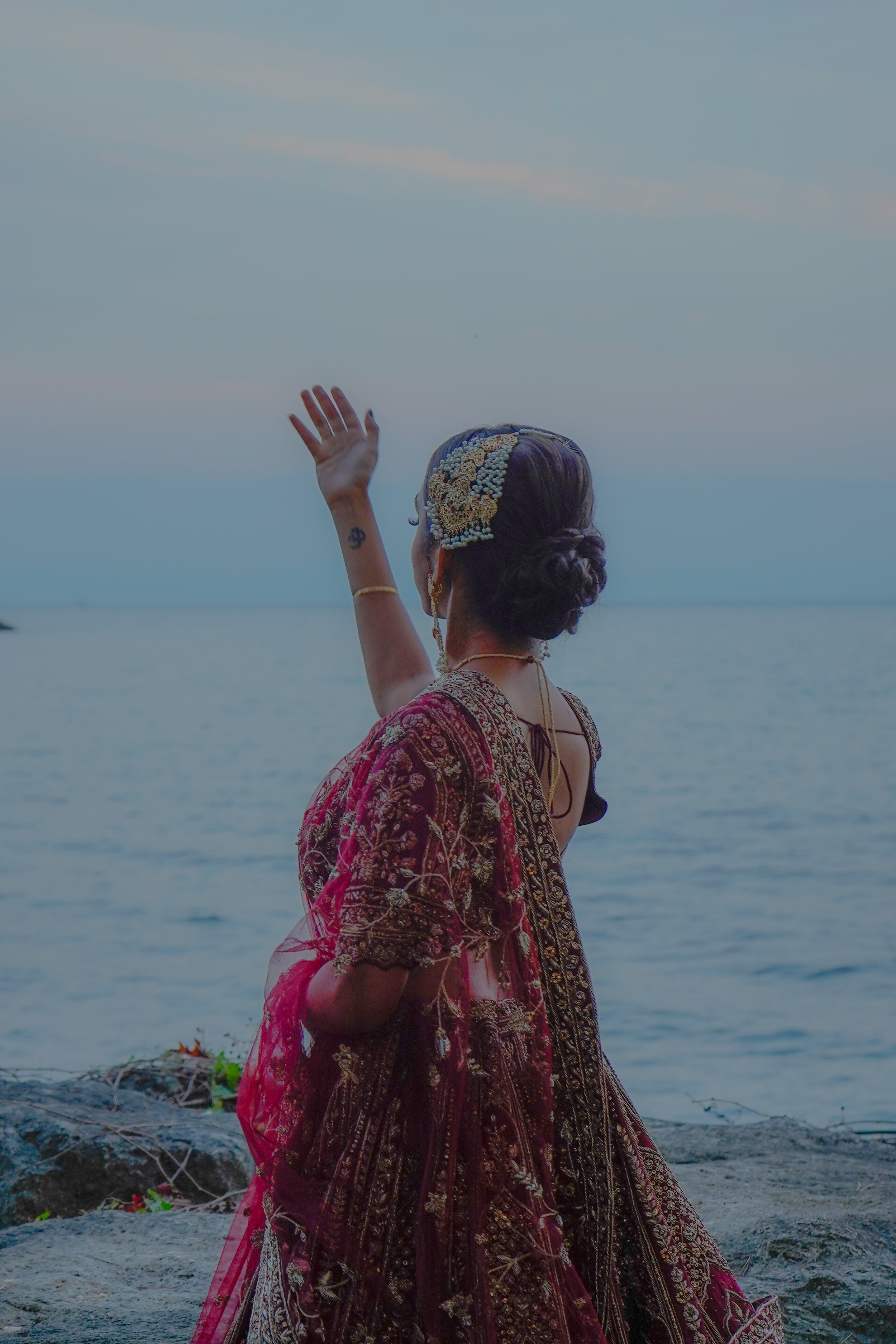 A woman dressed in traditional Indian clothing, with a floral headpiece, waving at the sea during sunset.