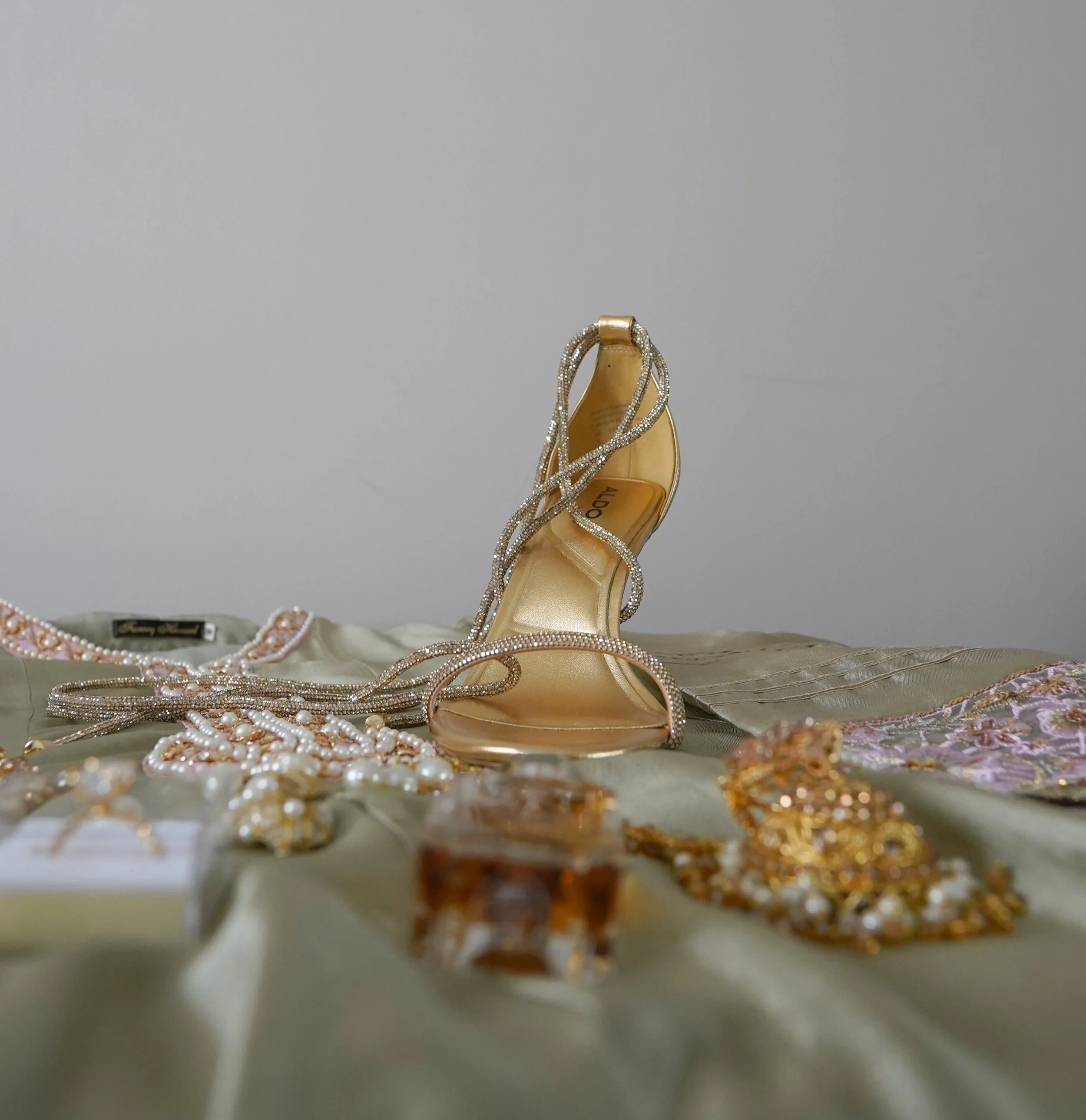 A gold high-heeled shoe with rhinestone straps, surrounded by pearl necklaces and jewelry, placed on a satin fabric surface.