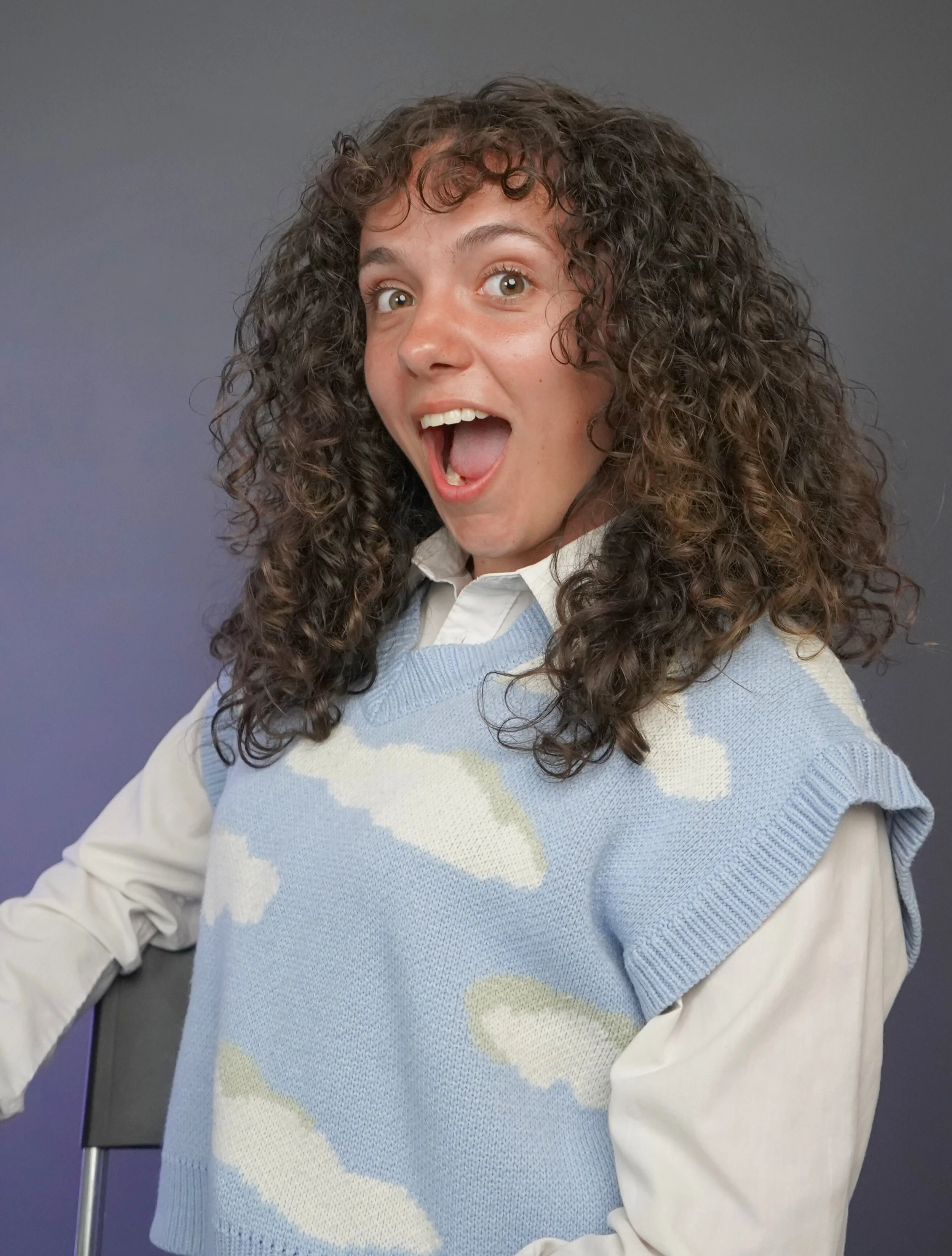 Excited woman with curly hair, wearing a white shirt and blue sweater with cloud patterns, sitting in front of a gray background.