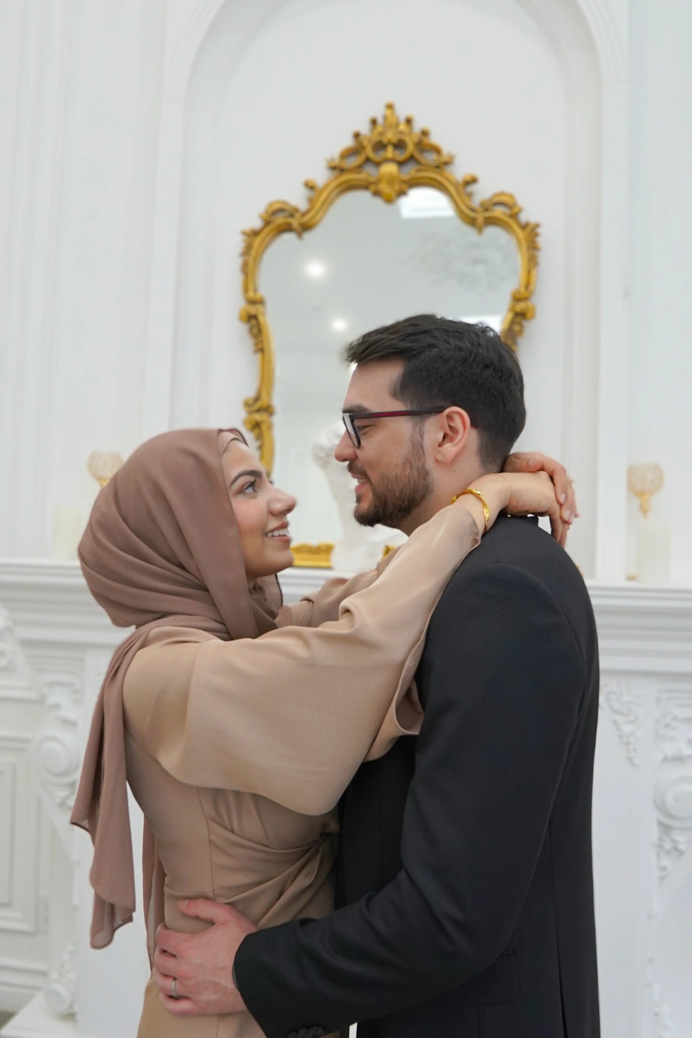 A couple embracing and looking into each other's eyes in an elegant room with gold accents and a large ornate mirror.