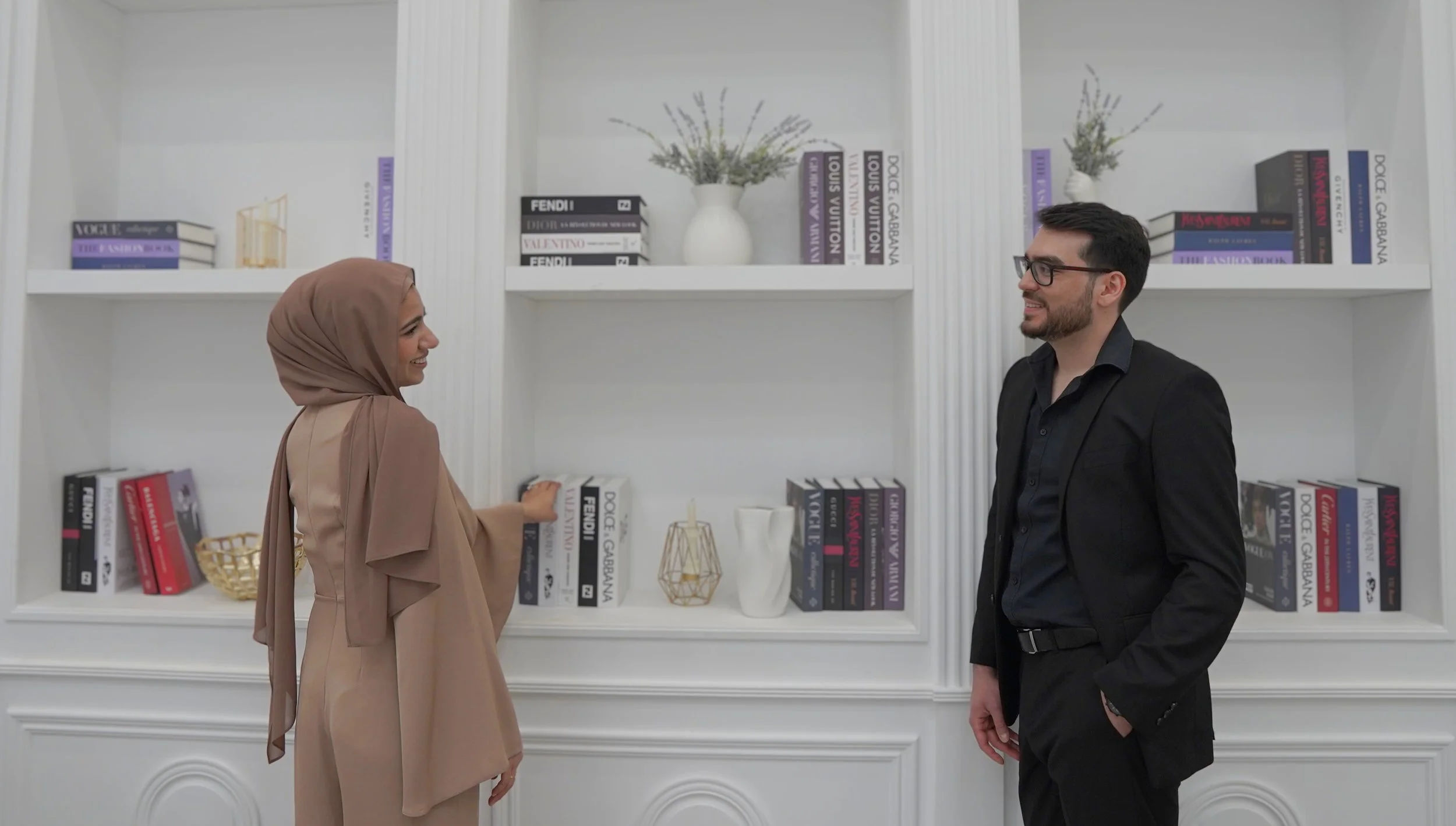 A woman in a beige hijab and matching robe smiling and talking to a man in a black suit and glasses, standing in front of white bookshelves filled with books and decorative items.