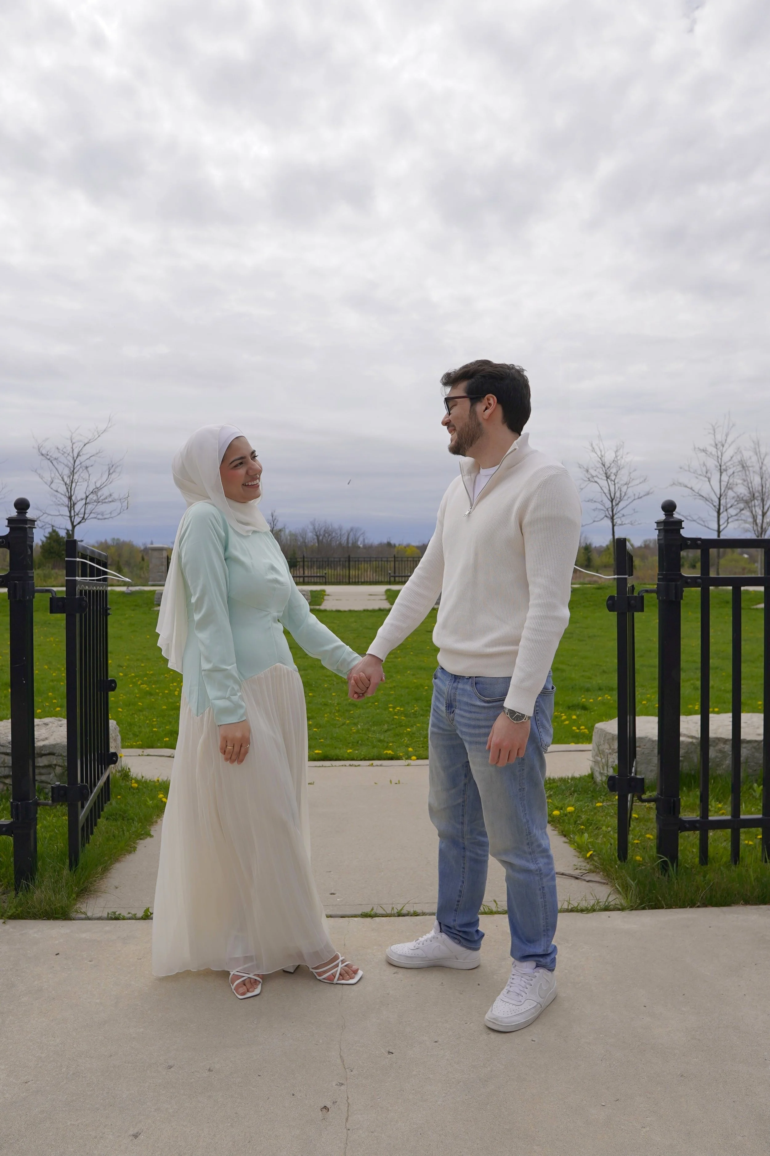 A couple standing outside on a cloudy day, holding hands and smiling at each other. The woman wears a light-colored dress and hijab, while the man wears a white sweater and jeans. They are near a black metal fence with a park and trees in the backgro