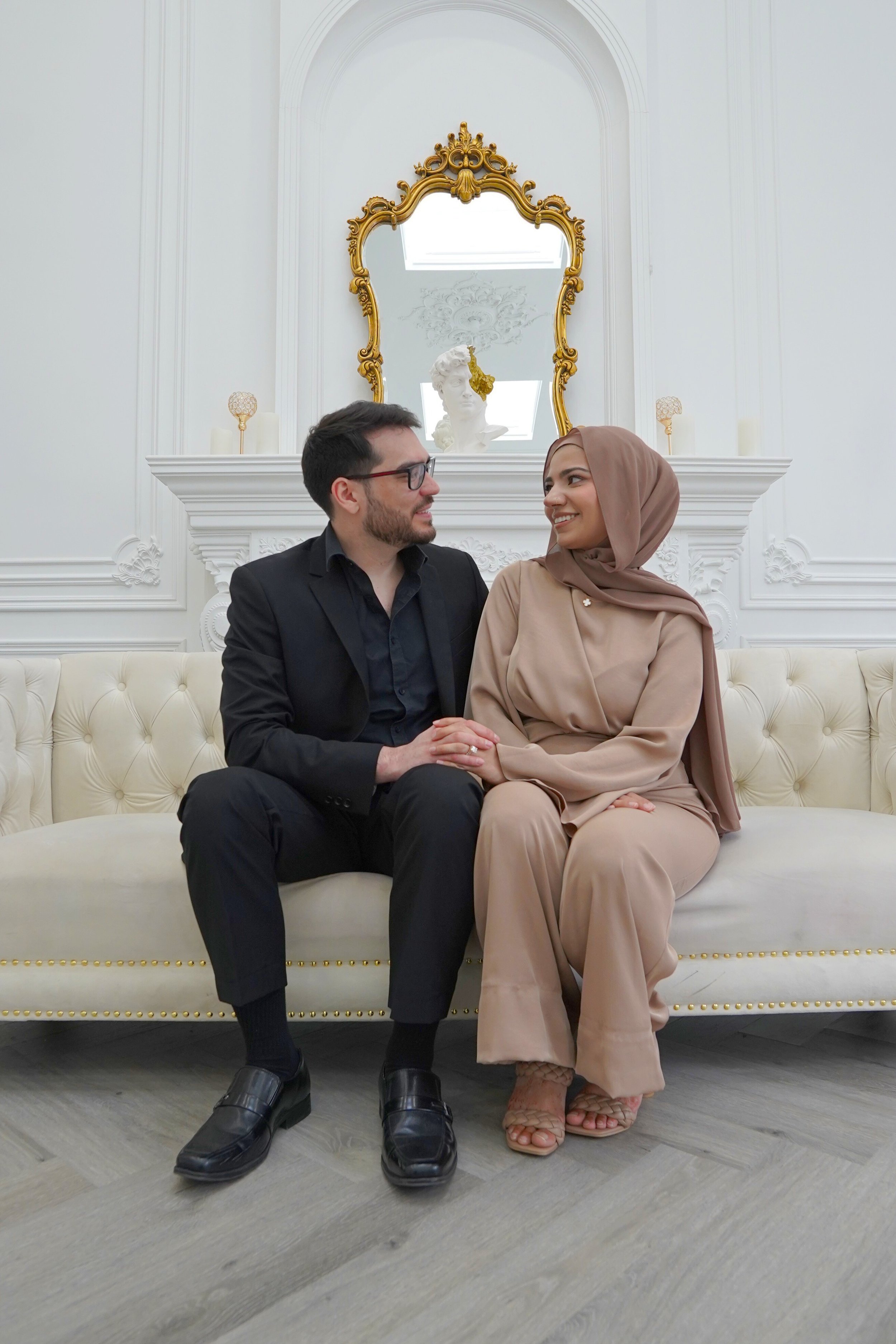 A man and woman sitting on a cream tufted sofa in an elegant room with white walls, gold accents, and a large ornate mirror.