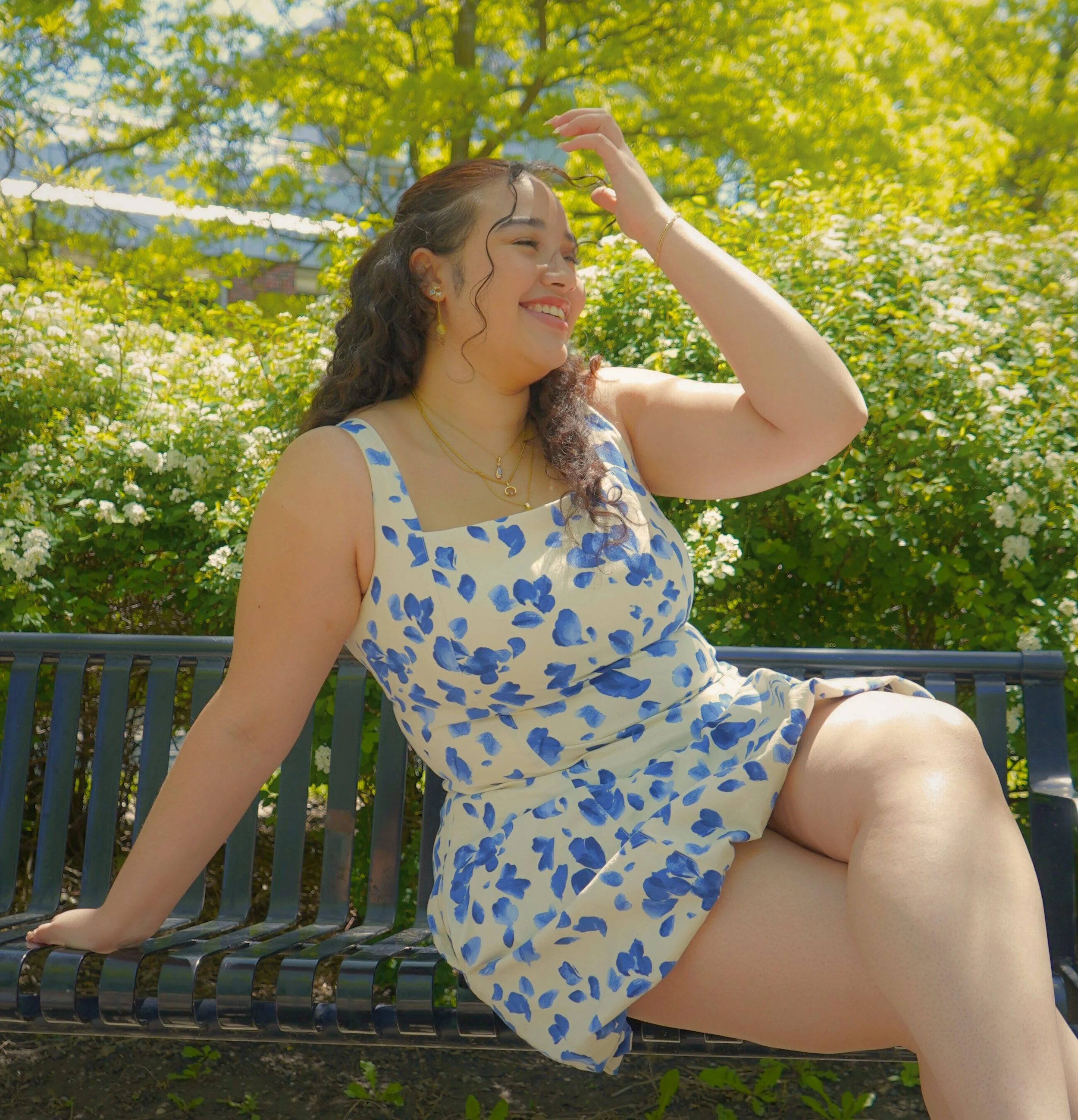 A woman with curly hair, wearing a white dress with blue watercolor-like spots, sitting on a black metal park bench surrounded by green bushes filled with white flowers, smiling and shading her eyes from the sun.