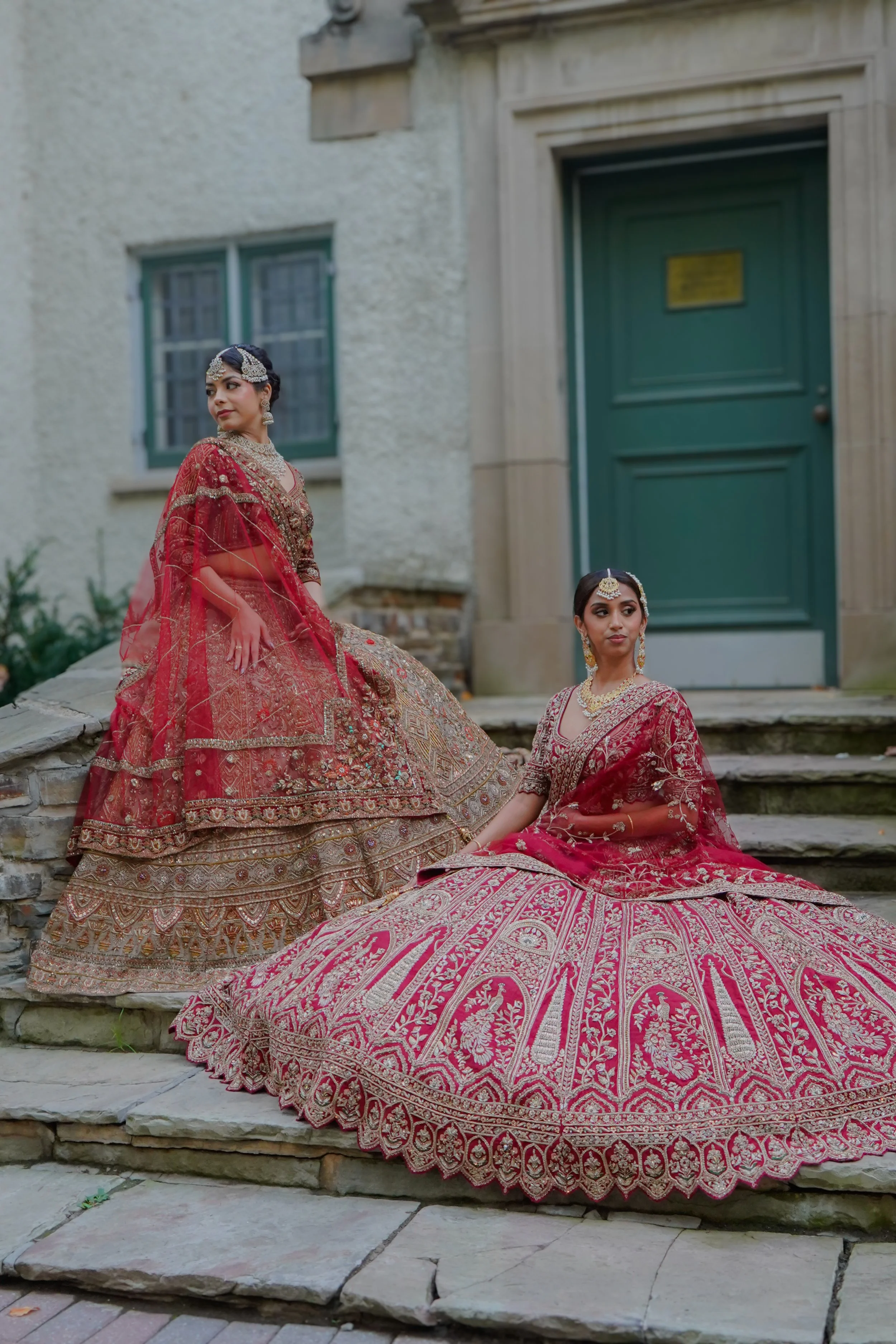 Two women dressed in traditional Indian bridal attire, with elaborate jewelry, sitting on stone steps outside a building with a green door.