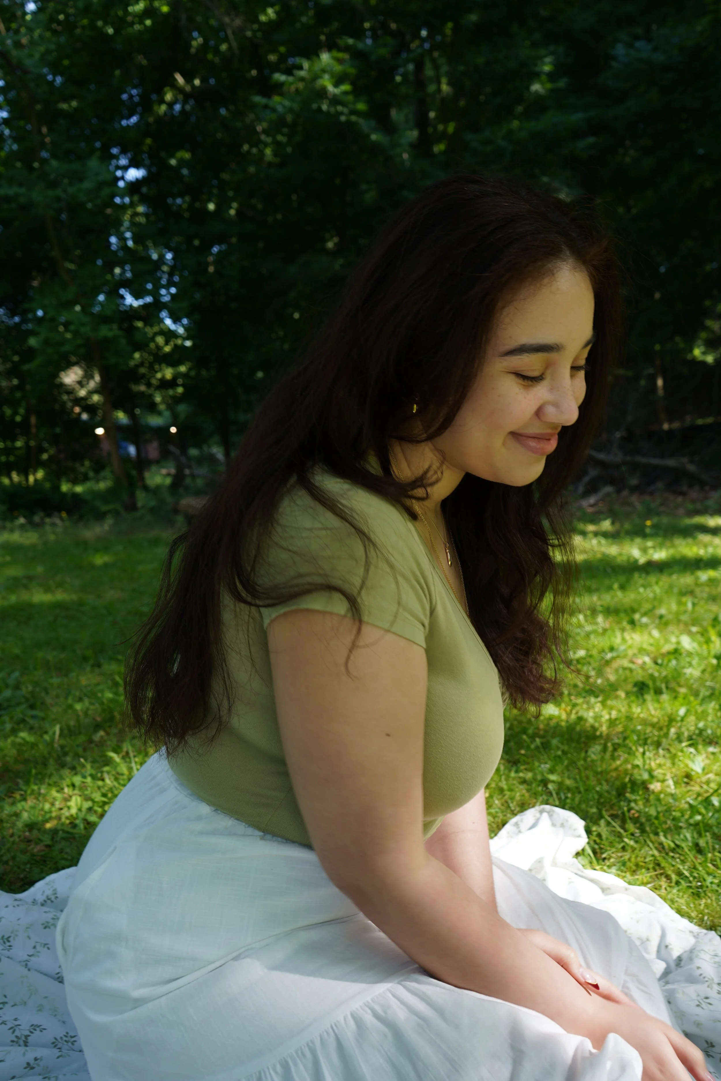 A woman sitting on grass with her eyes closed, smiling gently in an outdoor setting with trees and sunlight.