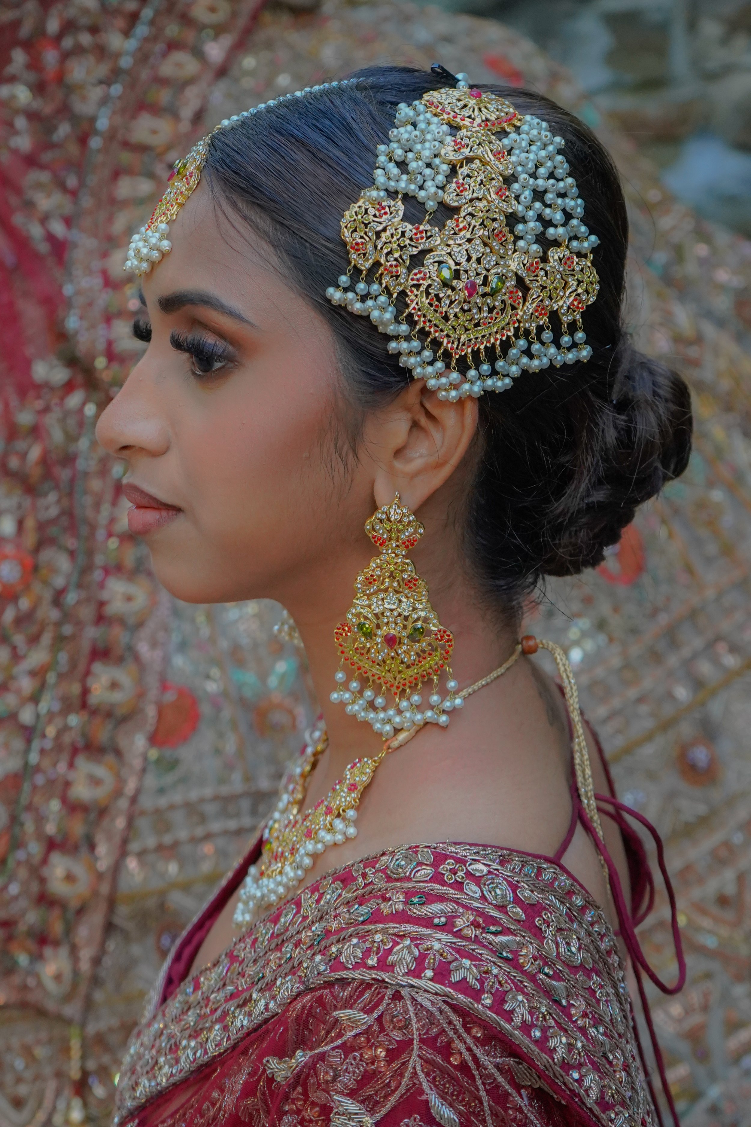 A woman in traditional Indian attire wearing elaborate gold and pearl jewelry, including earrings, a choker, and a maang tikka, with intricate embroidery on her maroon and gold dress, standing outdoors with blurred foliage in the background.