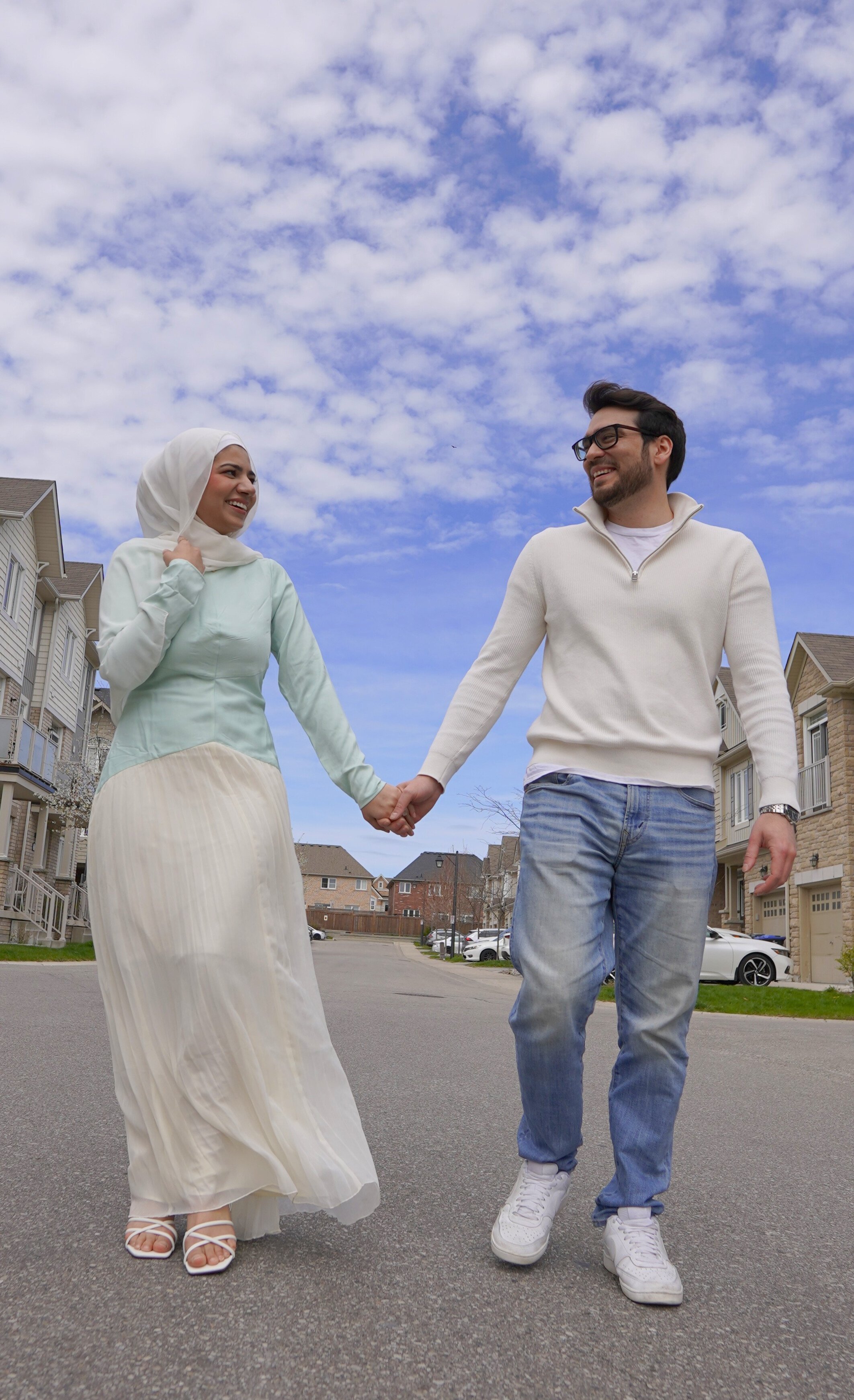 A couple walking hand-in-hand on a residential street, smiling and looking at each other, under a partly cloudy sky.