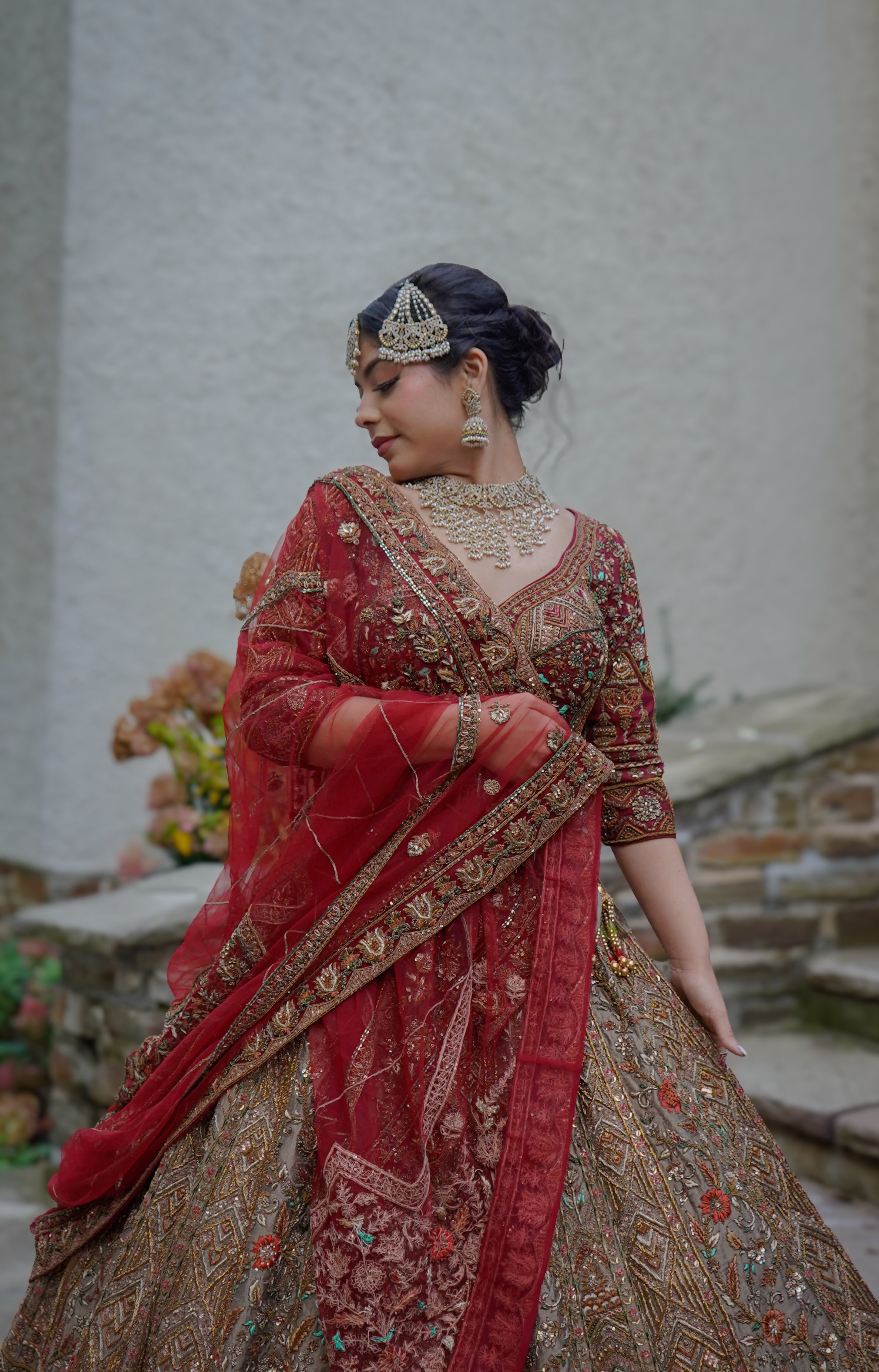 A woman dressed in traditional Indian bridal attire, wearing a heavily embroidered red and gold lehenga with jewelry, including a necklace, earrings, and a headpiece, standing against a neutral background with flowers in the background.