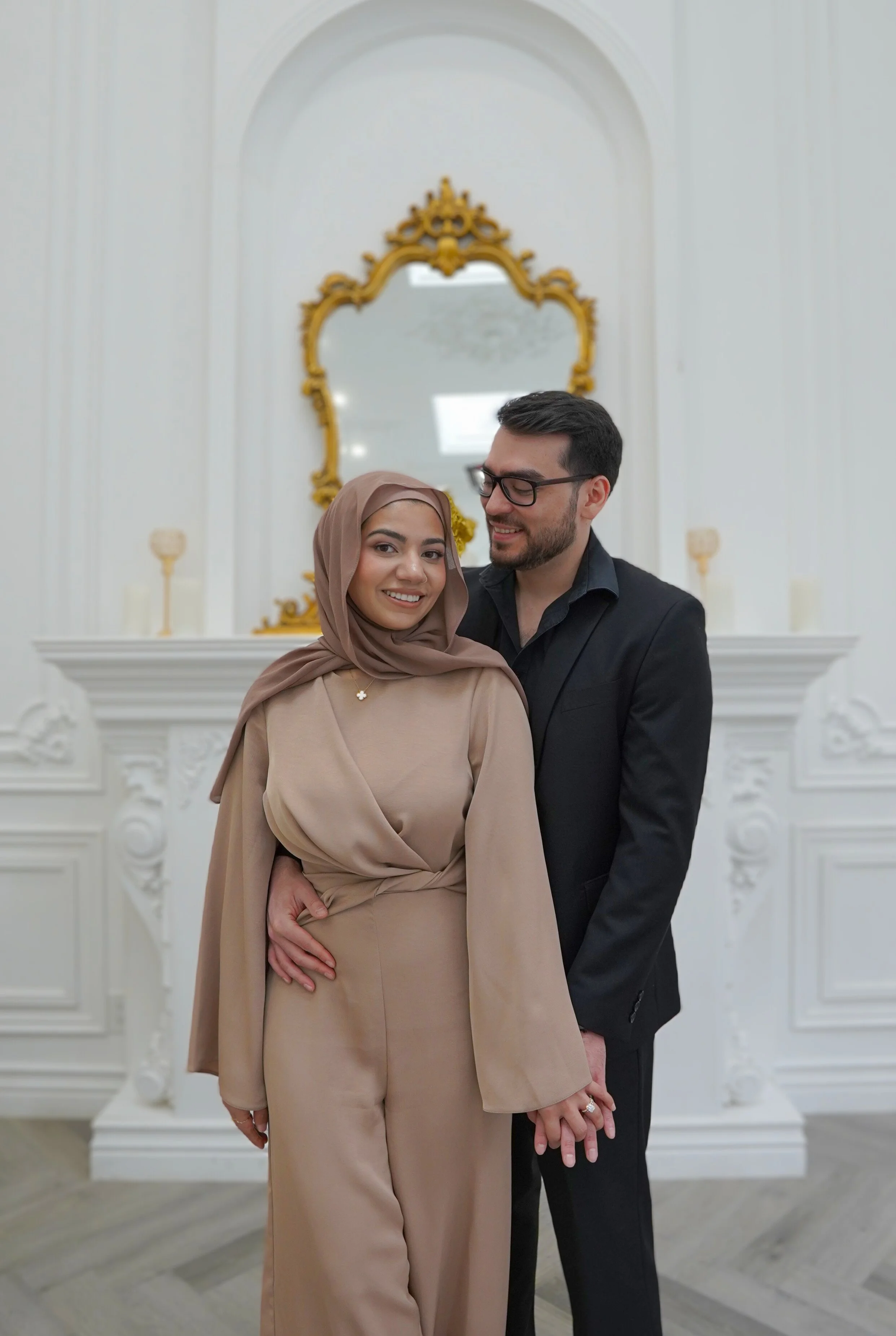 A couple dressed in elegant attire standing together in front of a white ornate fireplace with gold accents and a mirror, smiling and holding hands.