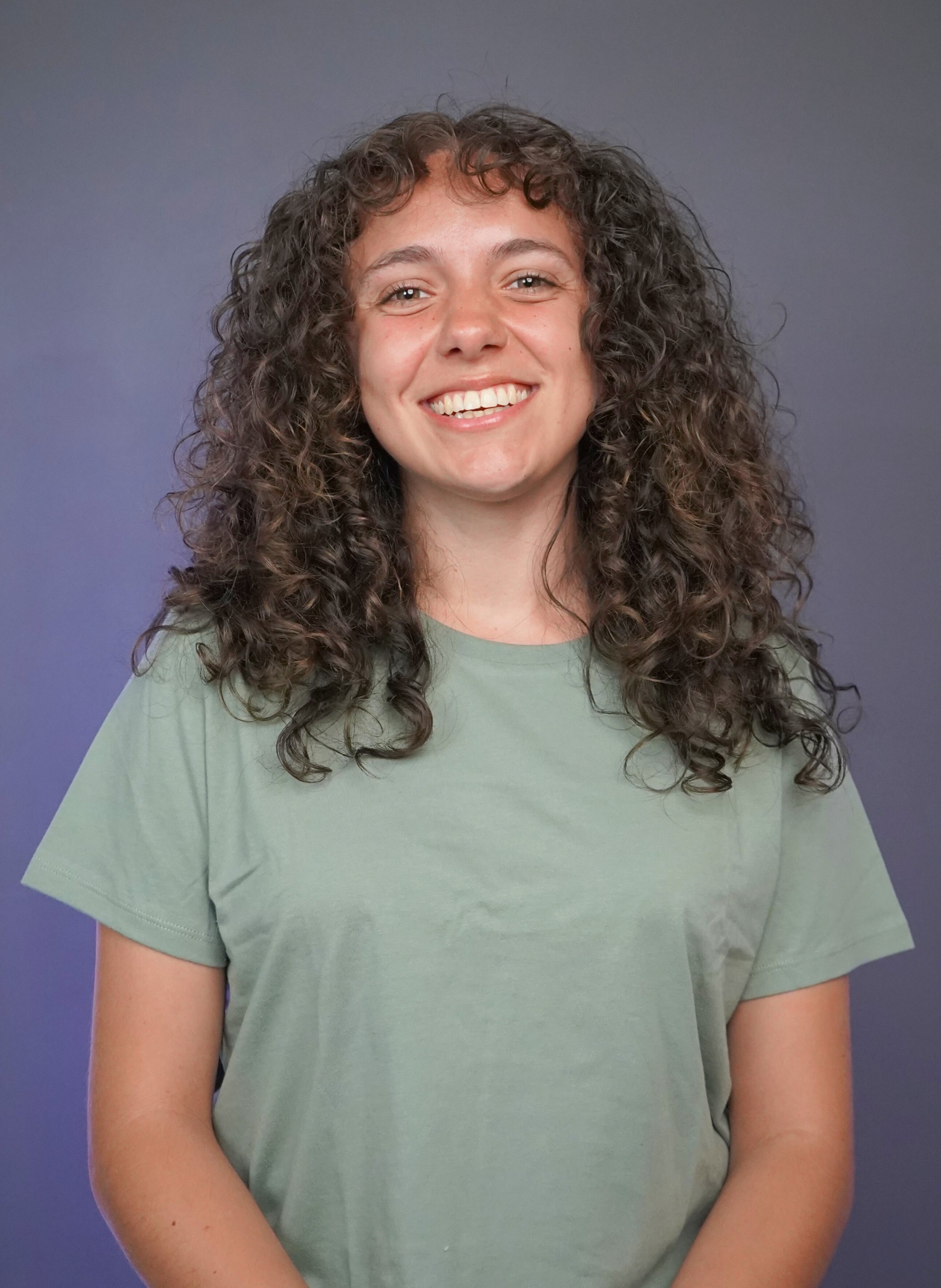 A woman with dark, curly hair smiling, wearing a light green t-shirt, standing against a dark gray background.