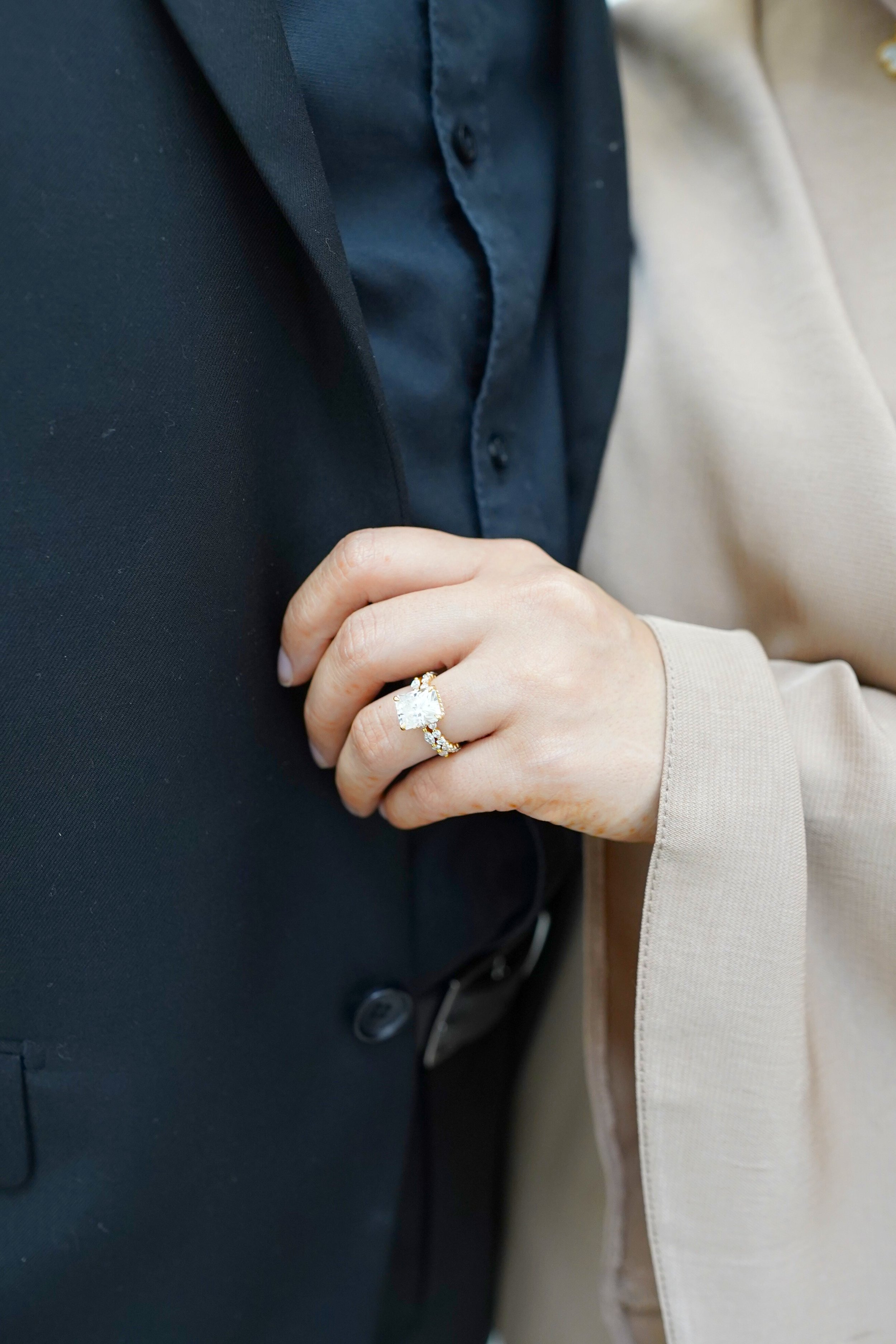 Close-up of a woman's hand with an engagement ring, touching a man's suit, with the woman wearing a light-colored blazer and the man in a dark suit.