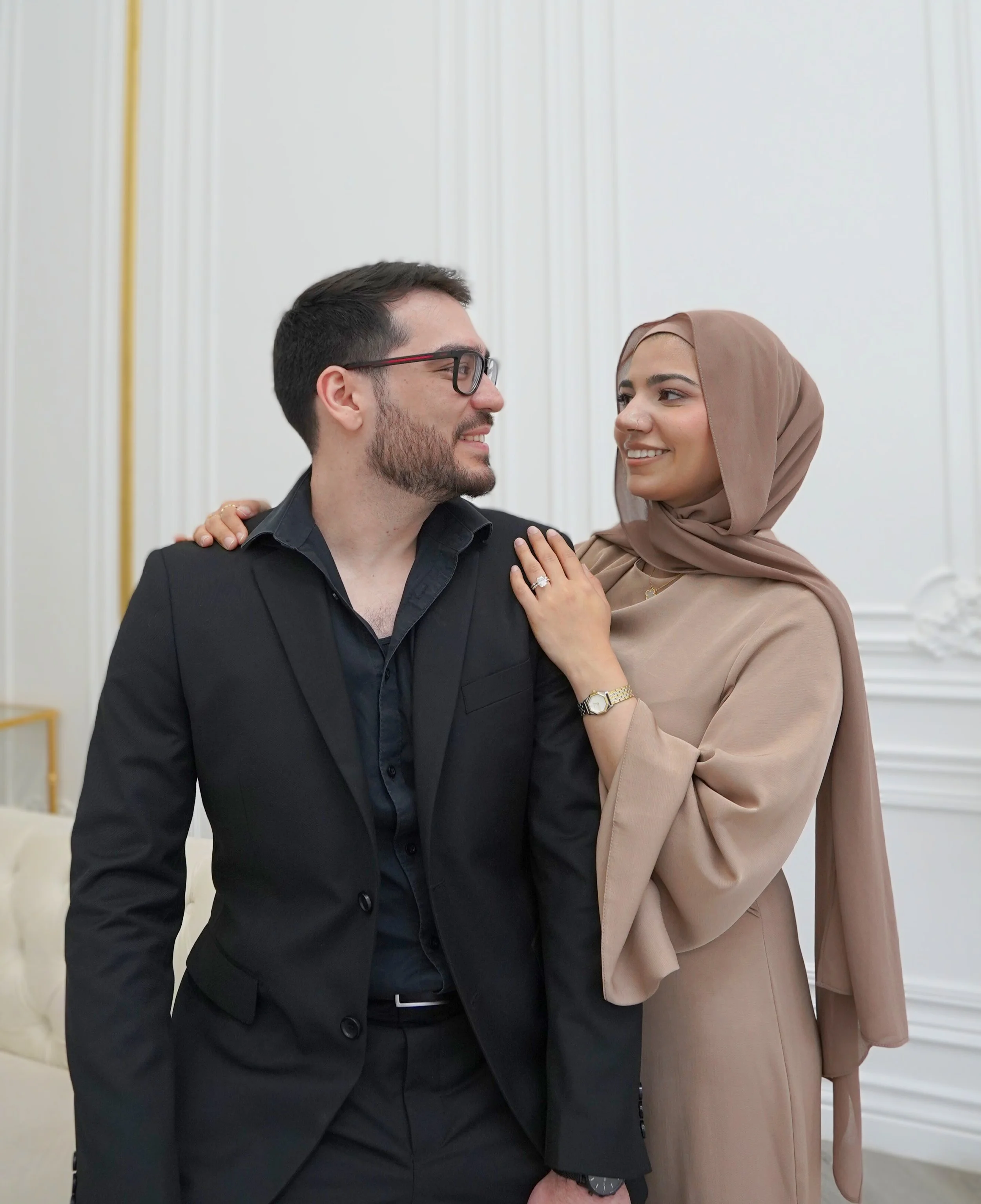 A man and woman stand close together, smiling at each other. The woman is wearing a beige hijab and dress, and the man is wearing glasses and a black suit. They are in a room with white walls and gold accents.