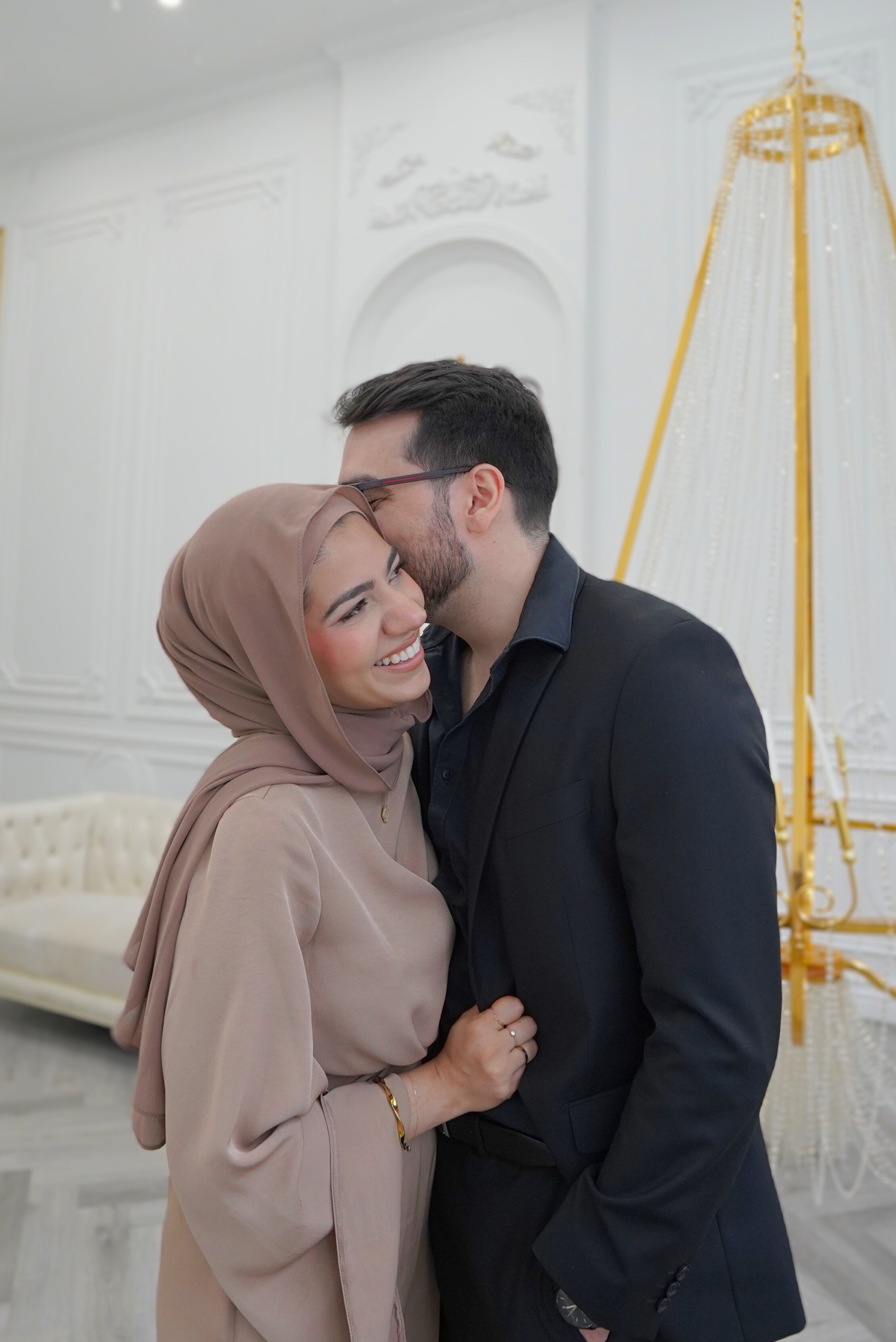 A smiling woman in a beige hijab and a black man in a suit share a joyful moment, with the man whispering into the woman's ear in a decorated indoor setting.
