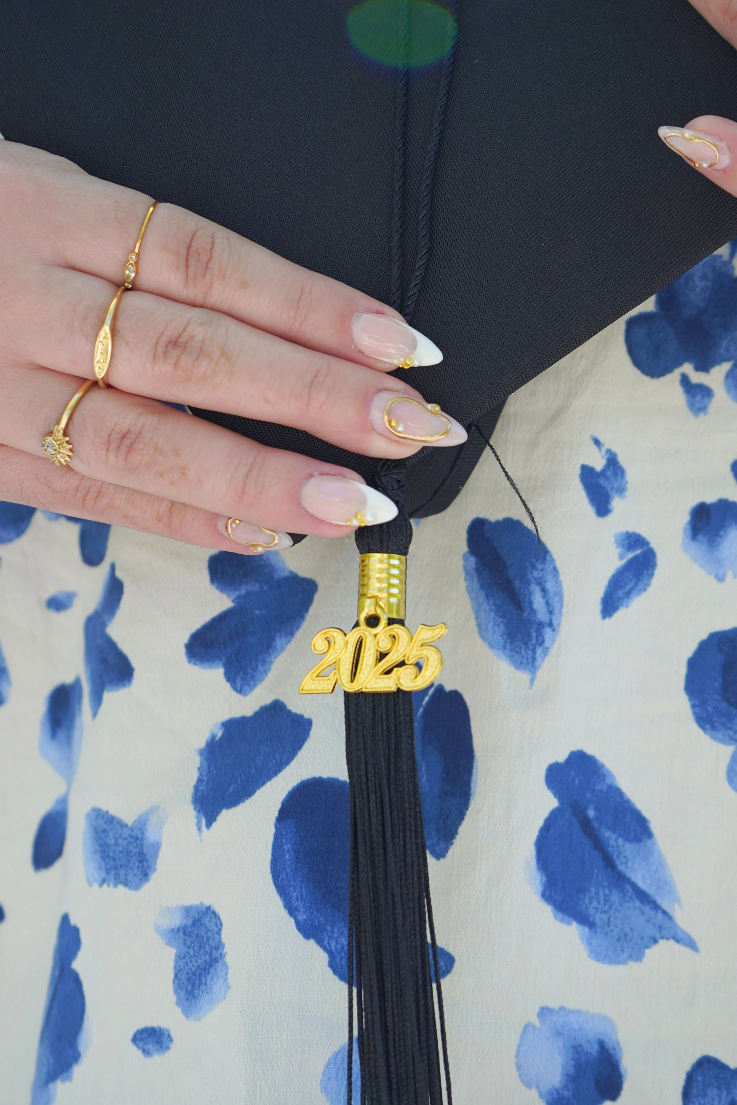 A person's hand with manicured nails holding a black graduation cap with a gold '2025' tassel, wearing several gold rings, positioned over a blue and white patterned fabric.