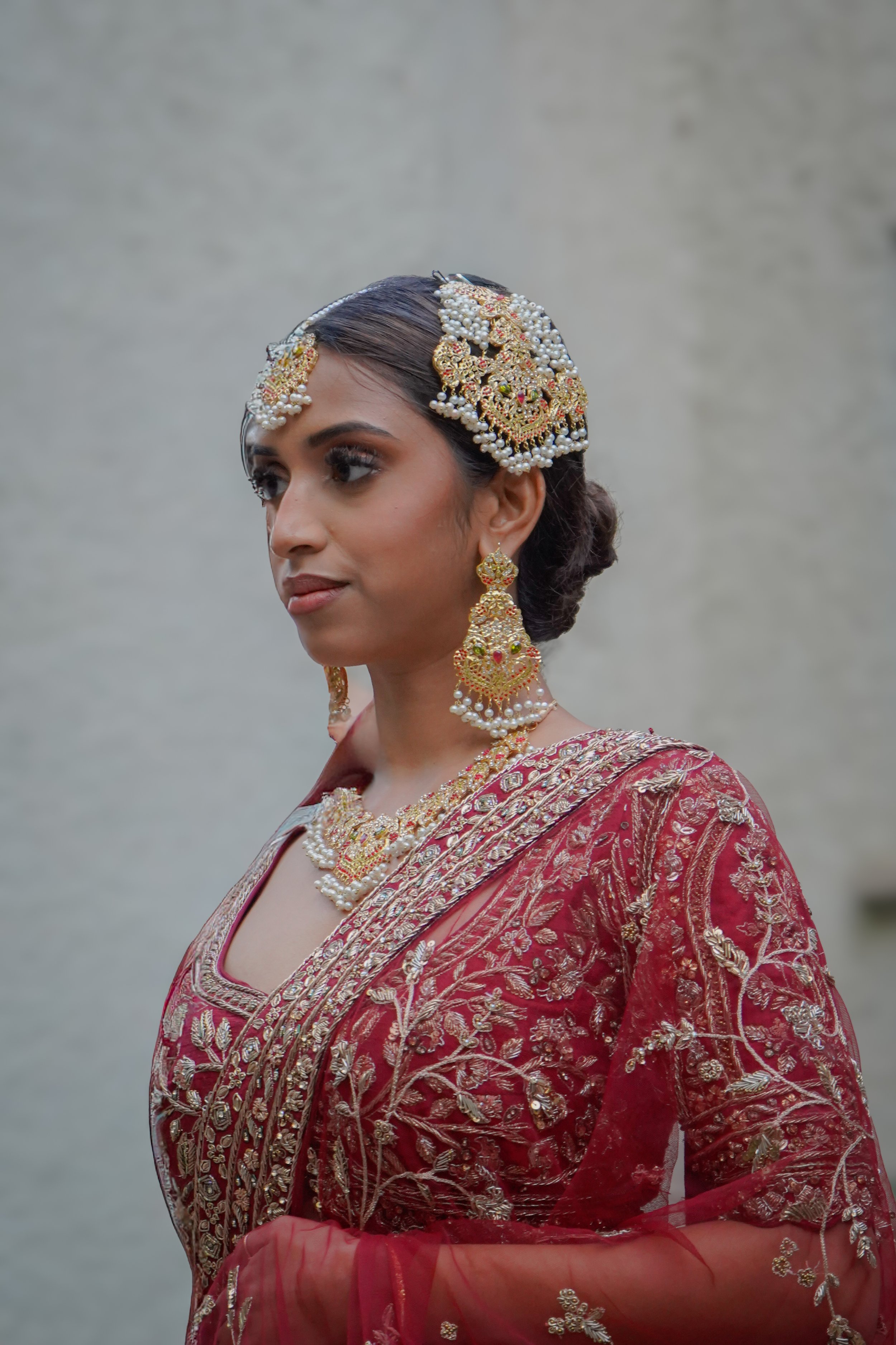 A woman dressed in traditional Indian bridal attire, wearing a red and gold embroidered saree with heavy gold jewelry, including earrings, necklace, and headpiece, against a plain background.
