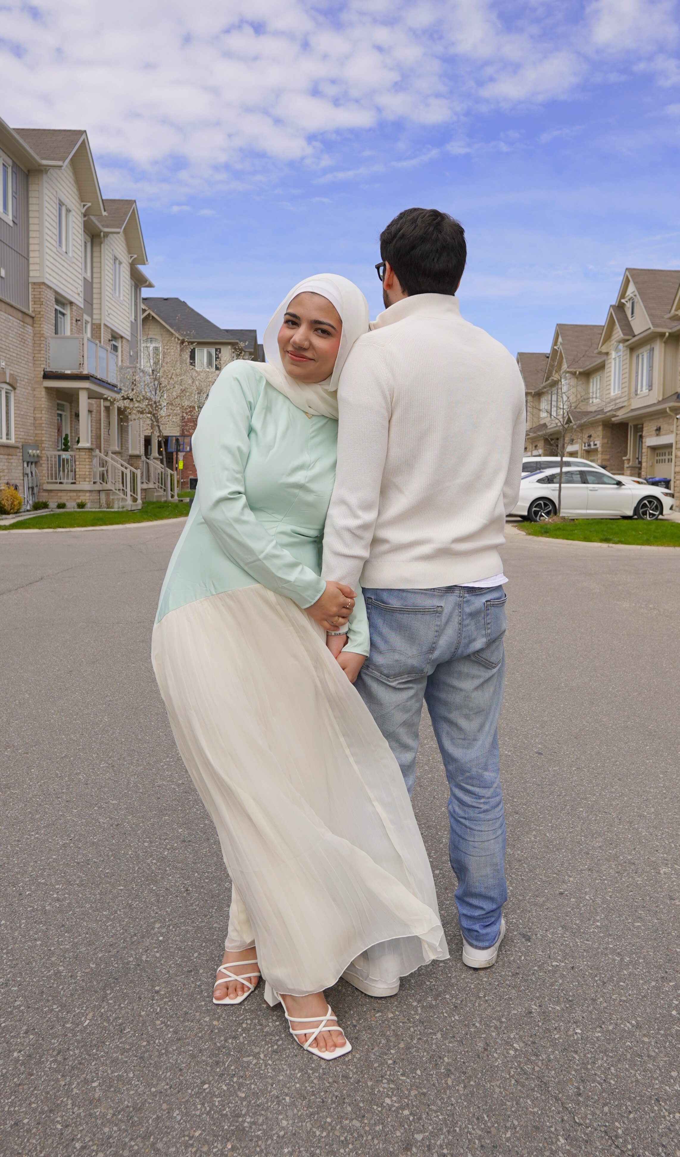 A woman wearing a white hijab and a light-colored dress standing next to a man in casual clothing, holding hands on a residential street with houses and parked cars, under a partly cloudy sky.