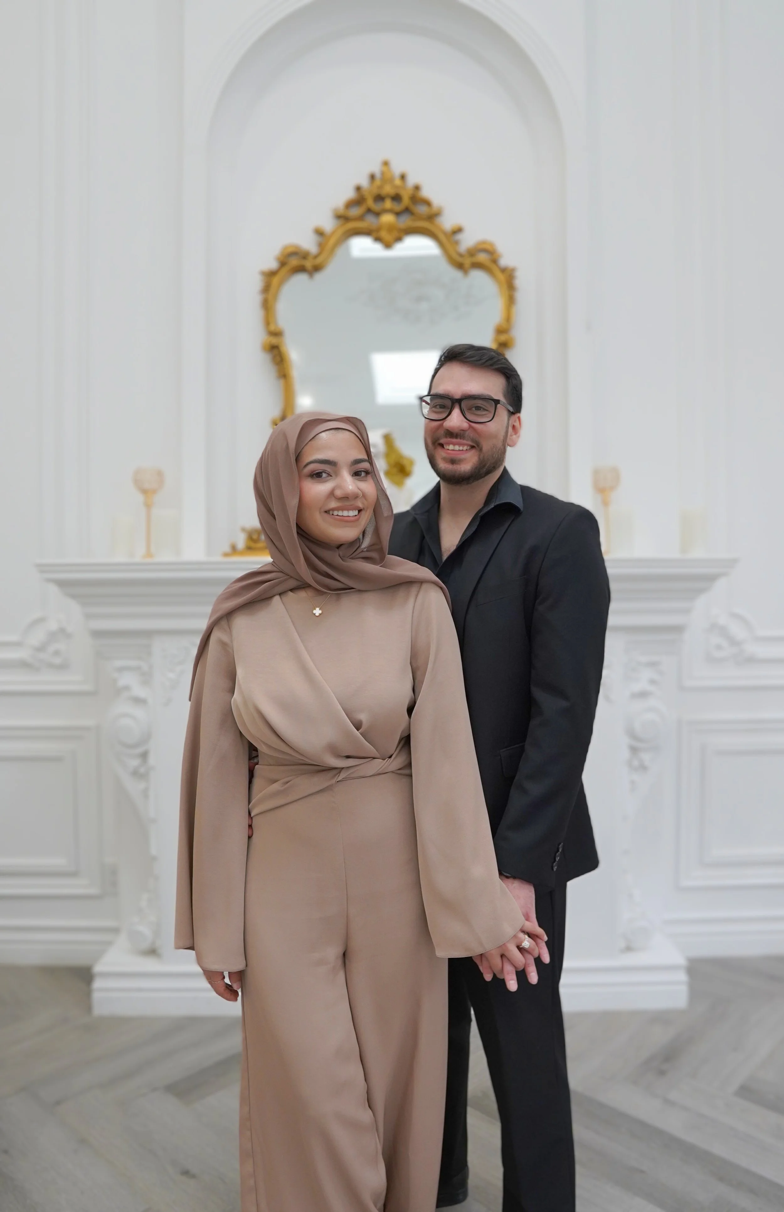A smiling couple standing inside a bright, ornate room with white walls and decorative gold accents, holding hands.