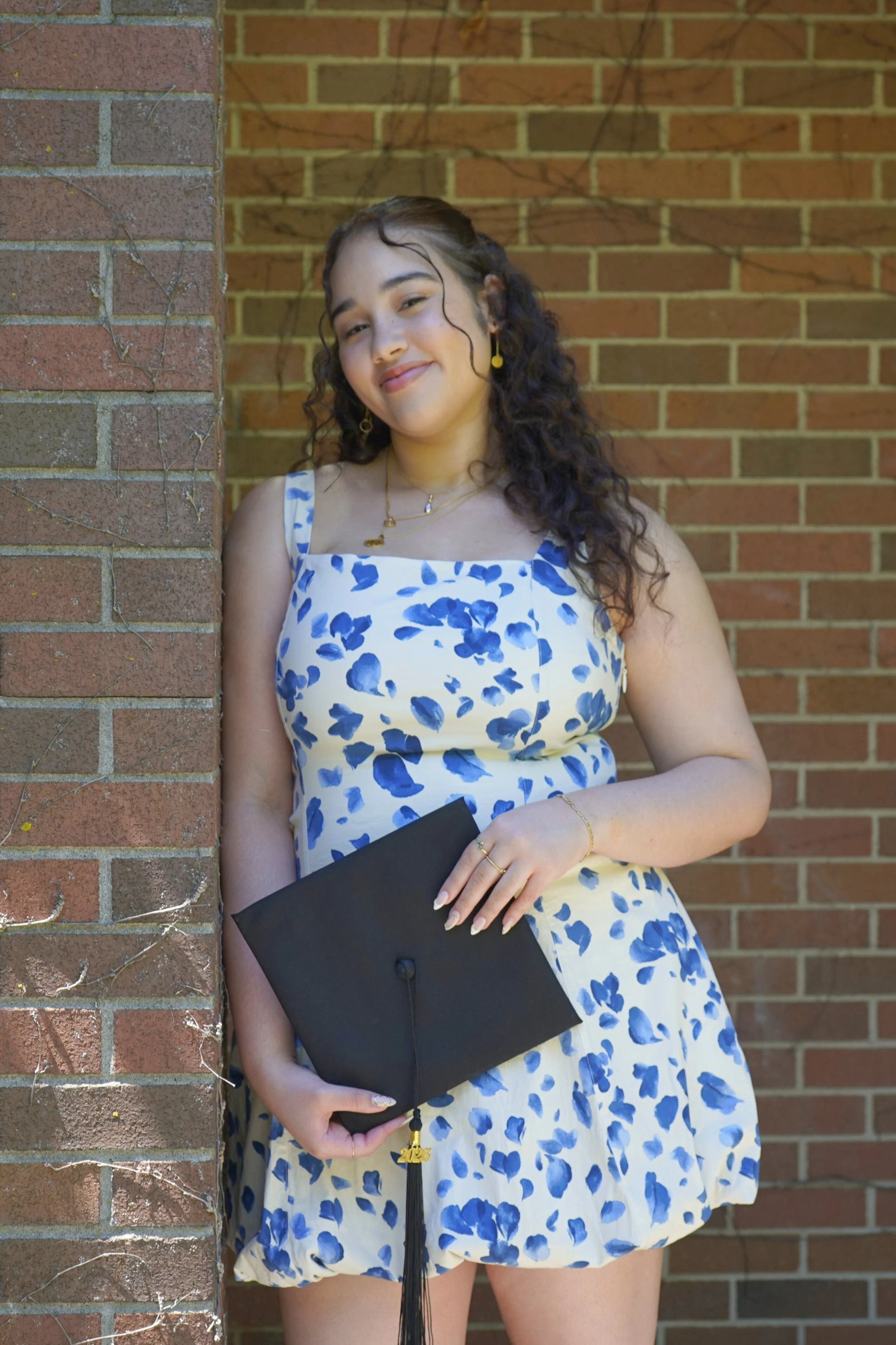 Young woman in a blue and white floral dress holding a black graduation cap, standing against a brick wall, smiling.