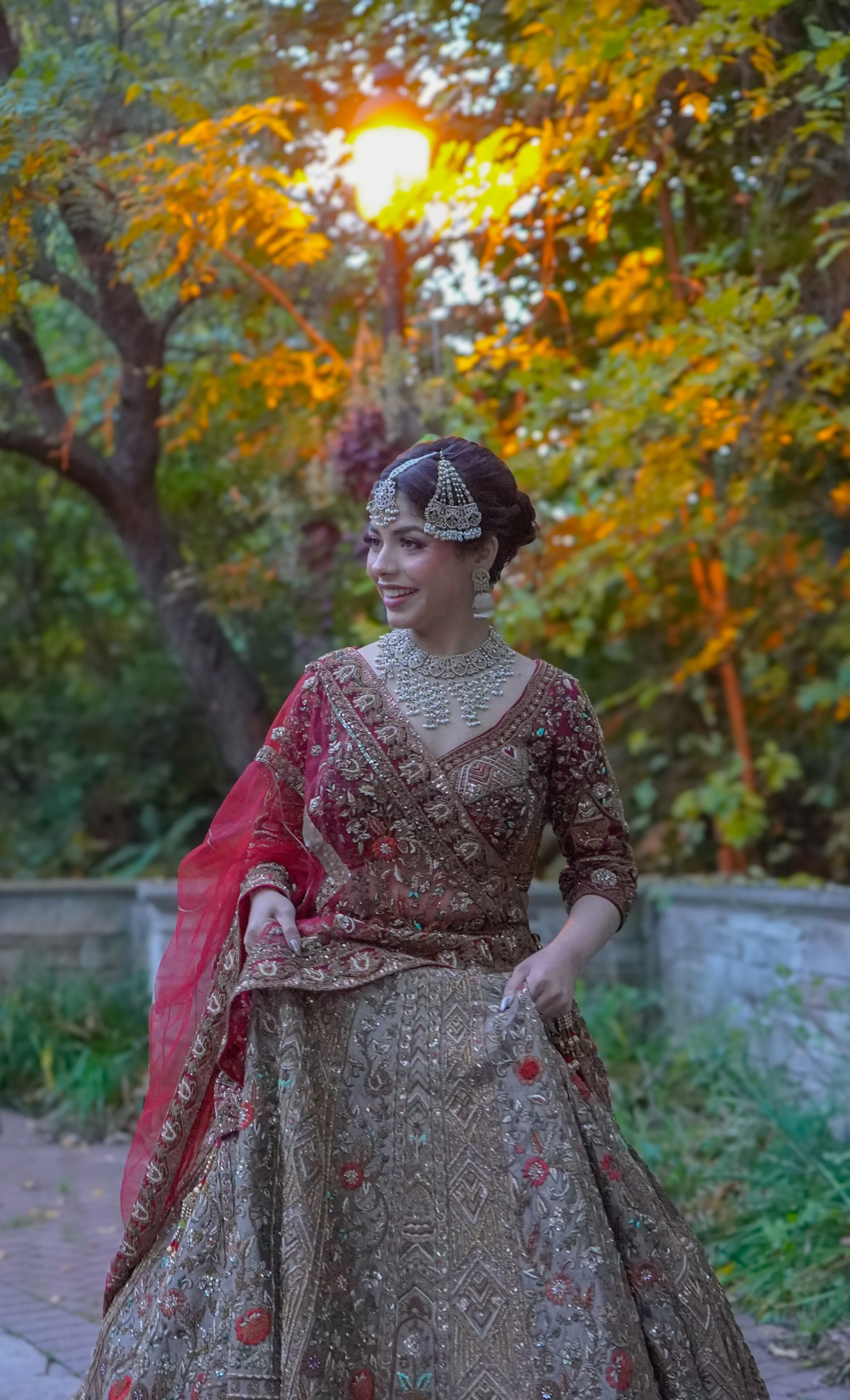 A woman dressed in traditional Indian bridal attire, wearing an intricately embroidered red and gold lehenga, with heavy jewelry including a necklace, earrings, and a headpiece, standing outdoors against a backdrop of trees with autumn-colored leaves