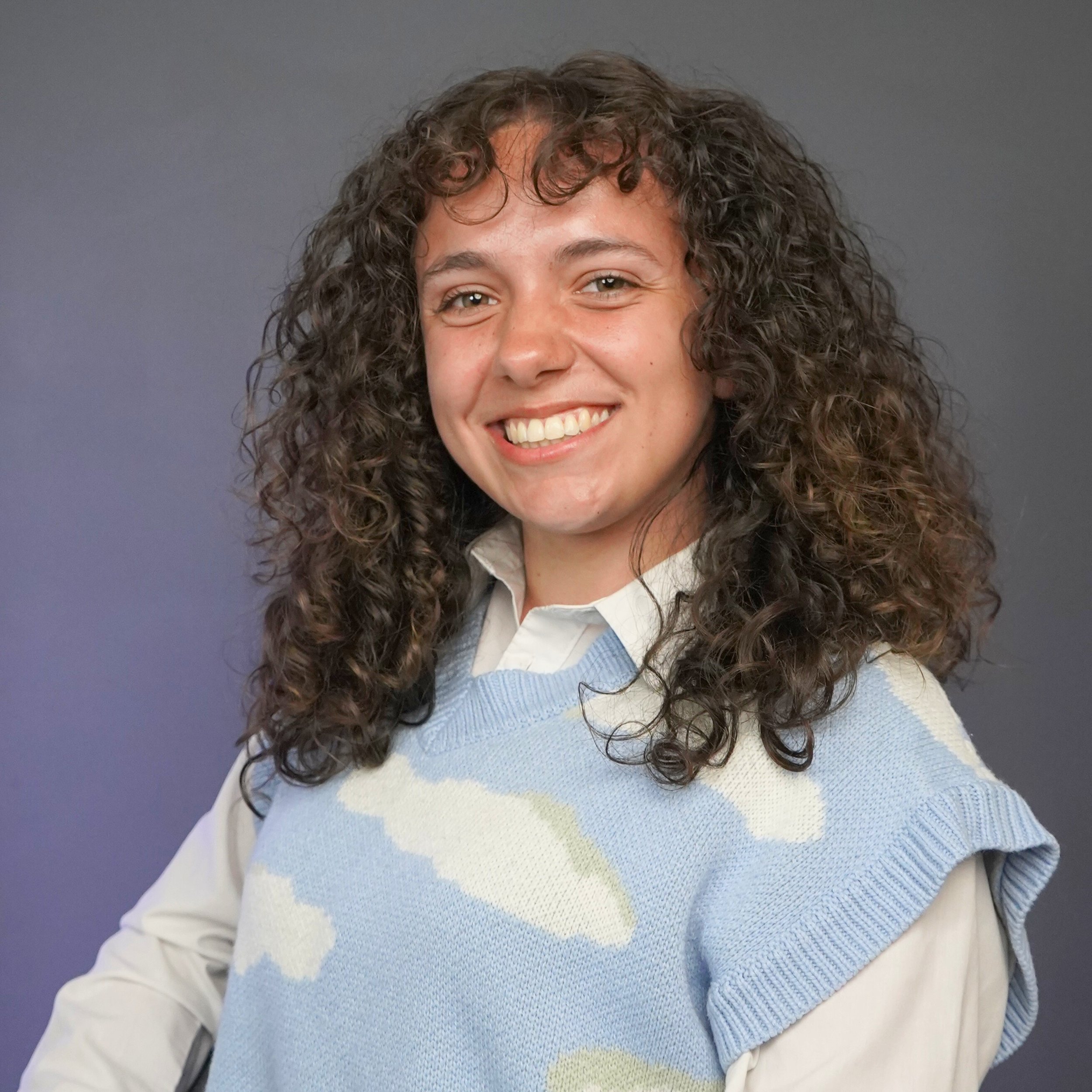 A young woman with long curly brown hair smiling, wearing a layered outfit with a white shirt and a light blue sweater with white cloud patterns, standing against a plain dark background.