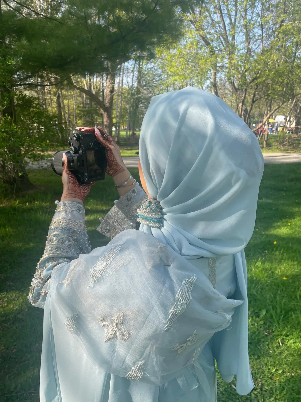 A woman in a light blue traditional dress with intricate embroidery is holding a camera outdoors in a park with green grass and trees.