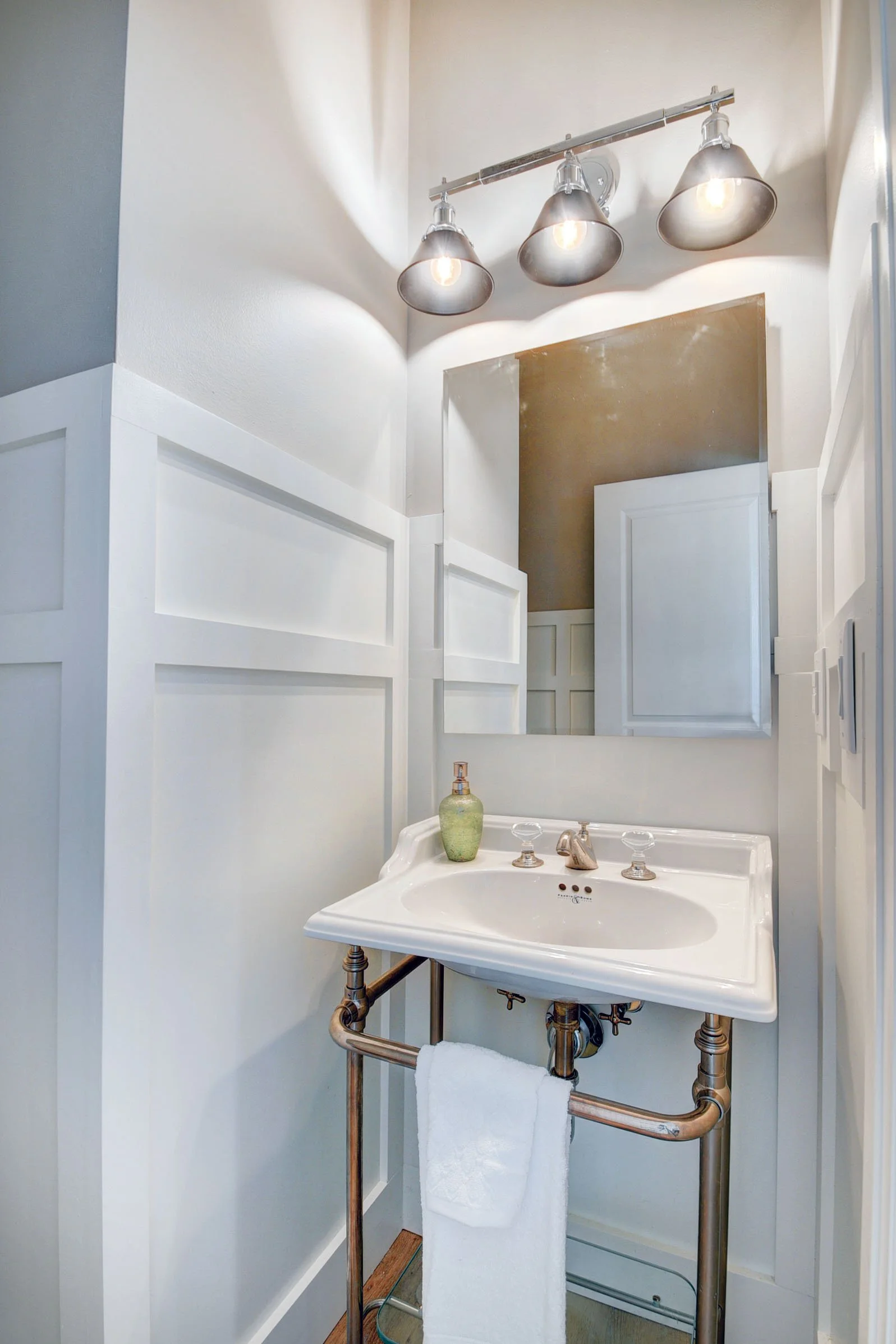 Small bathroom with white paneled walls, a vintage porcelain sink with exposed metal piping, and modern industrial-style three-light fixture with metal shades.