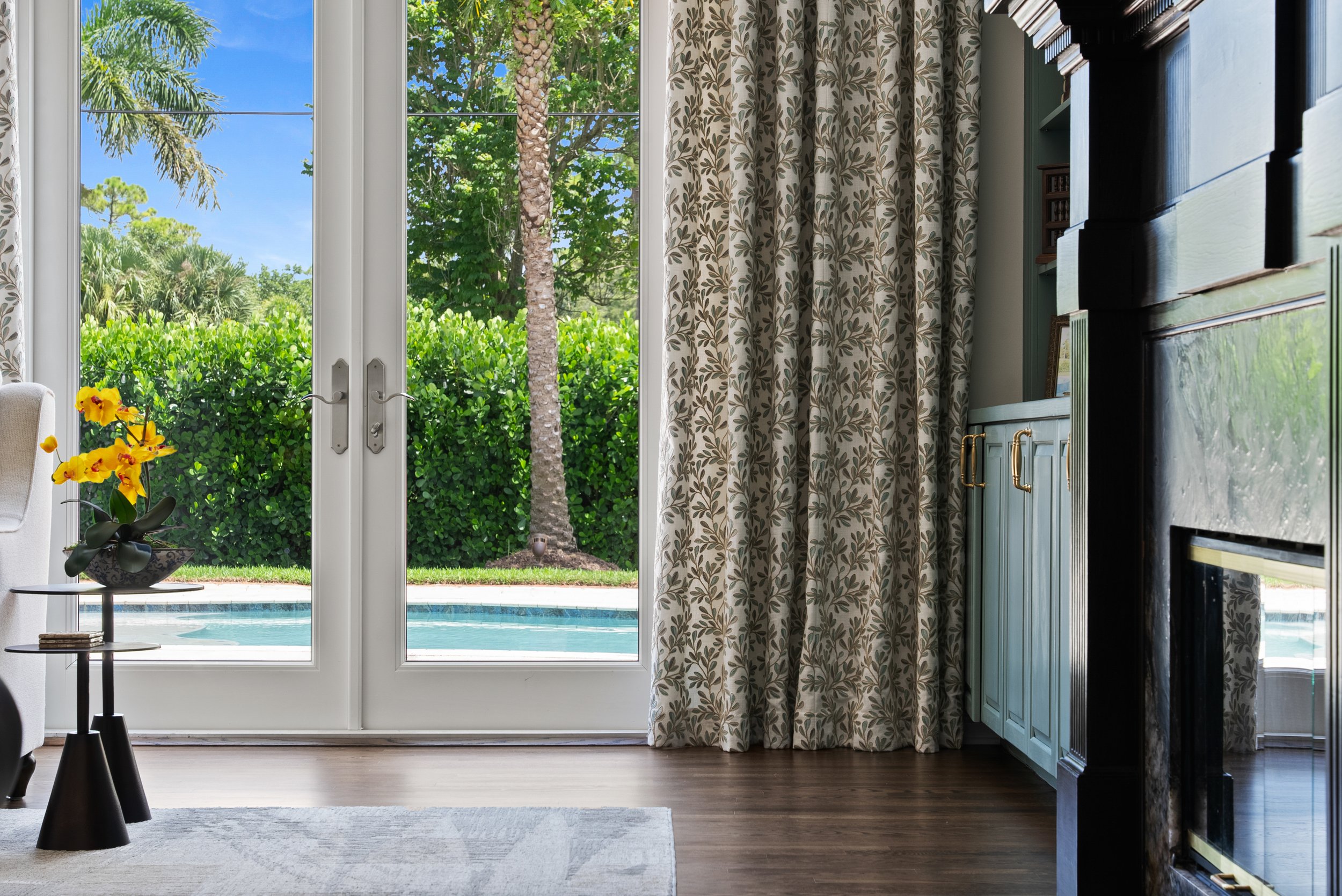 Interior view of a living room with glass doors leading outside to a garden and pool, floral curtains, a side table with yellow orchids, and hardwood floors.