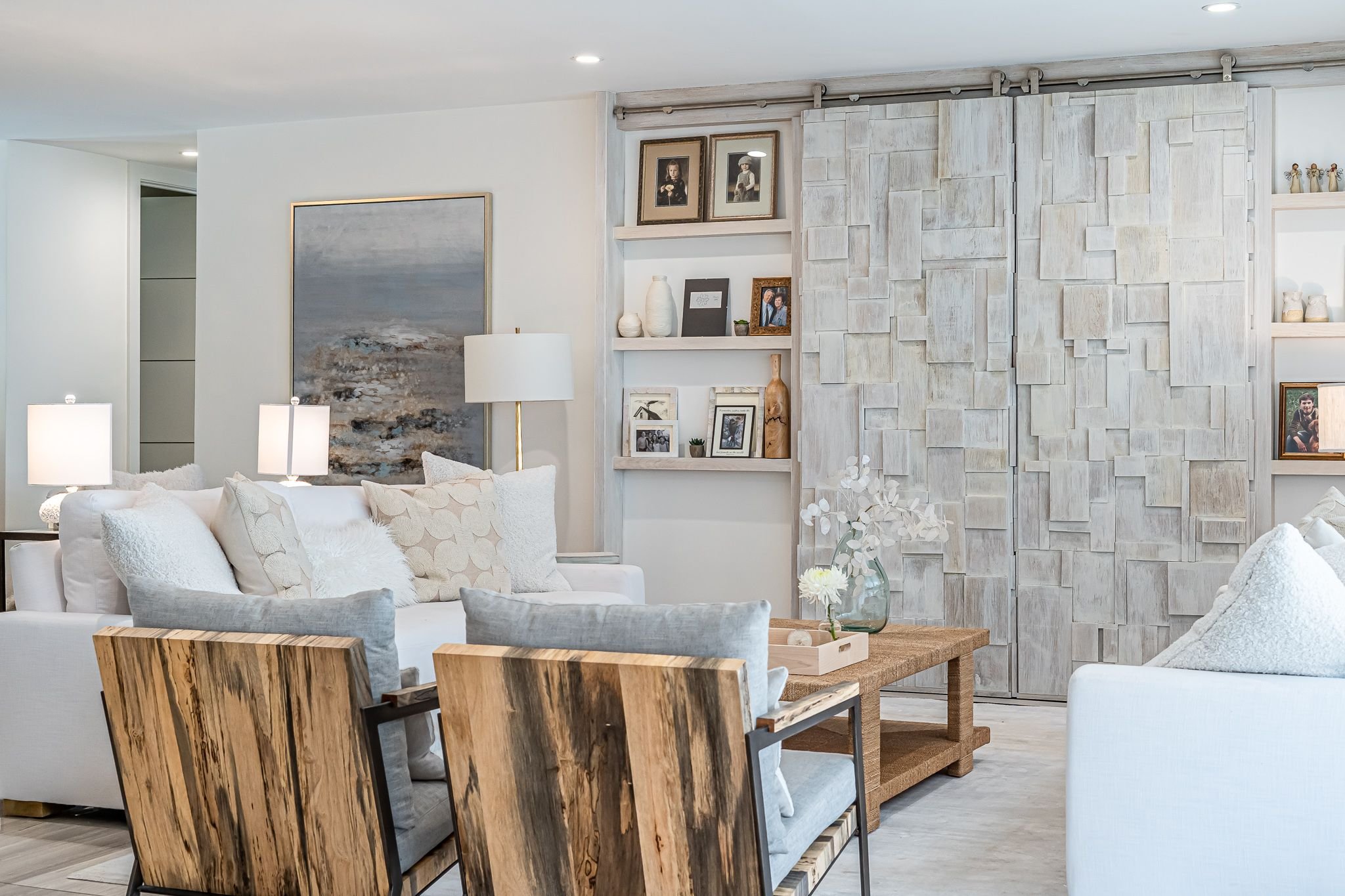 Living room with white sofas, wooden chairs, a coffee table with flowers, framed wall art, built-in shelves with photos and decor, and a textured sliding door.