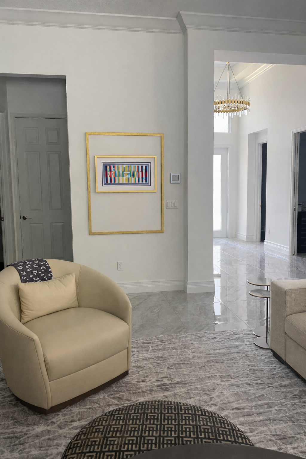 Living room with cream-colored armchair, modern abstract artwork framed in gold on white wall, marble flooring, and entryway leading to bright hallway with chandelier.