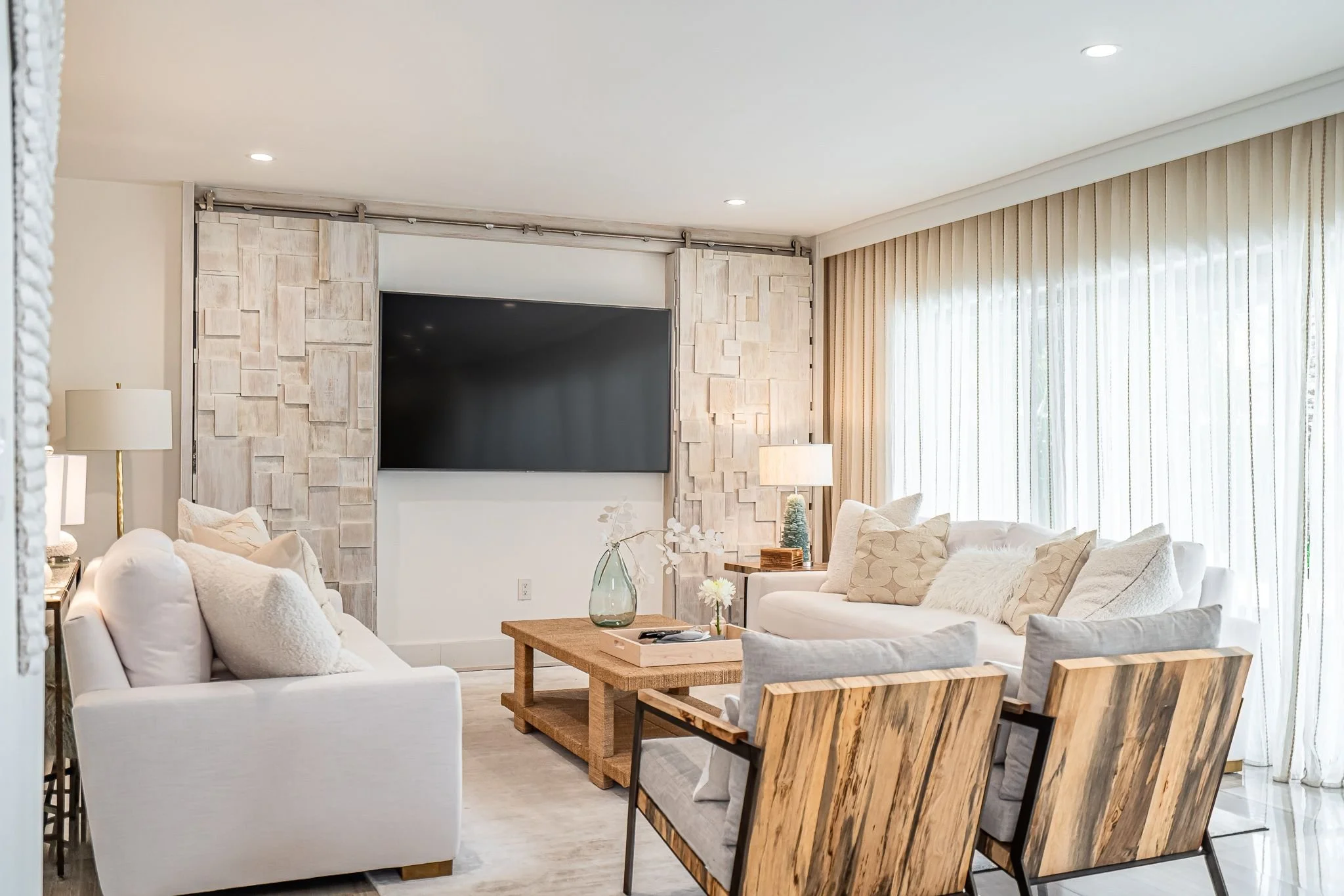 Bright living room with white sofas and armchairs, light wood coffee table, large windows with sheer curtains, decorative pillows, and a wall-mounted flat screen TV.