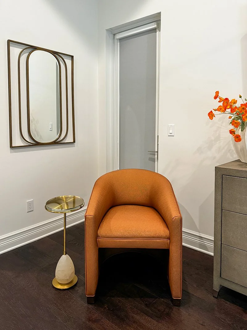 A cozy corner with an orange upholstered armchair, a small gold and marble side table, a modern gray dresser with orange flowers in a white vase, a white wall with a decorative mirror, and a door in the background.