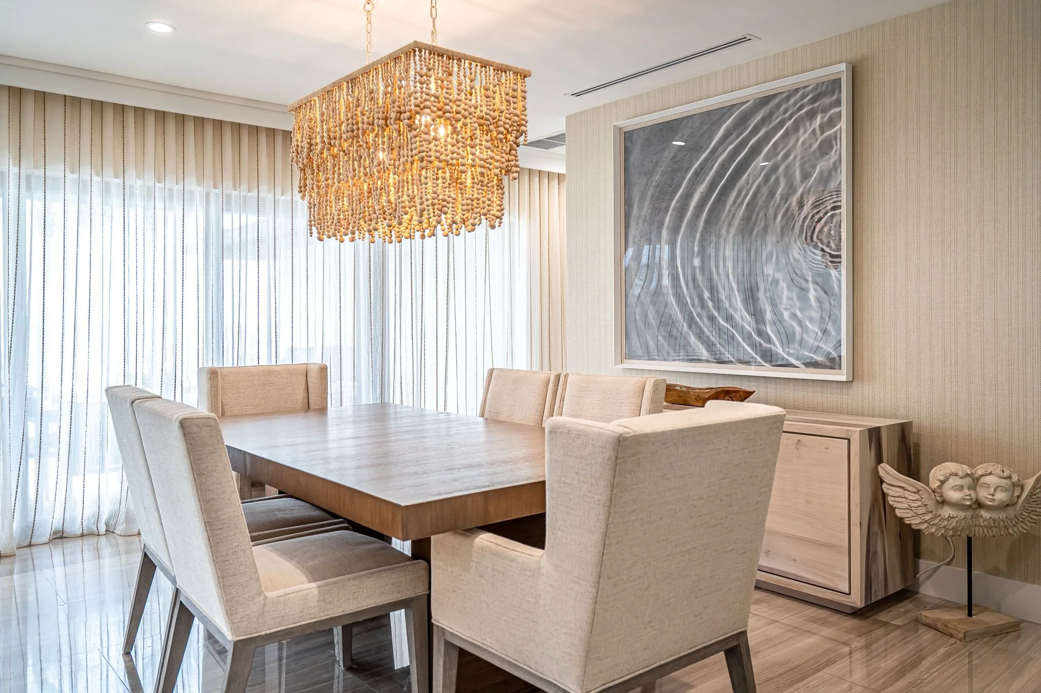 Modern dining room with a wooden table and six beige upholstered chairs, large window with sheer curtains, a colorful chandelier, a framed abstract picture on the wall, and a wooden sideboard with decorative angel sculptures.