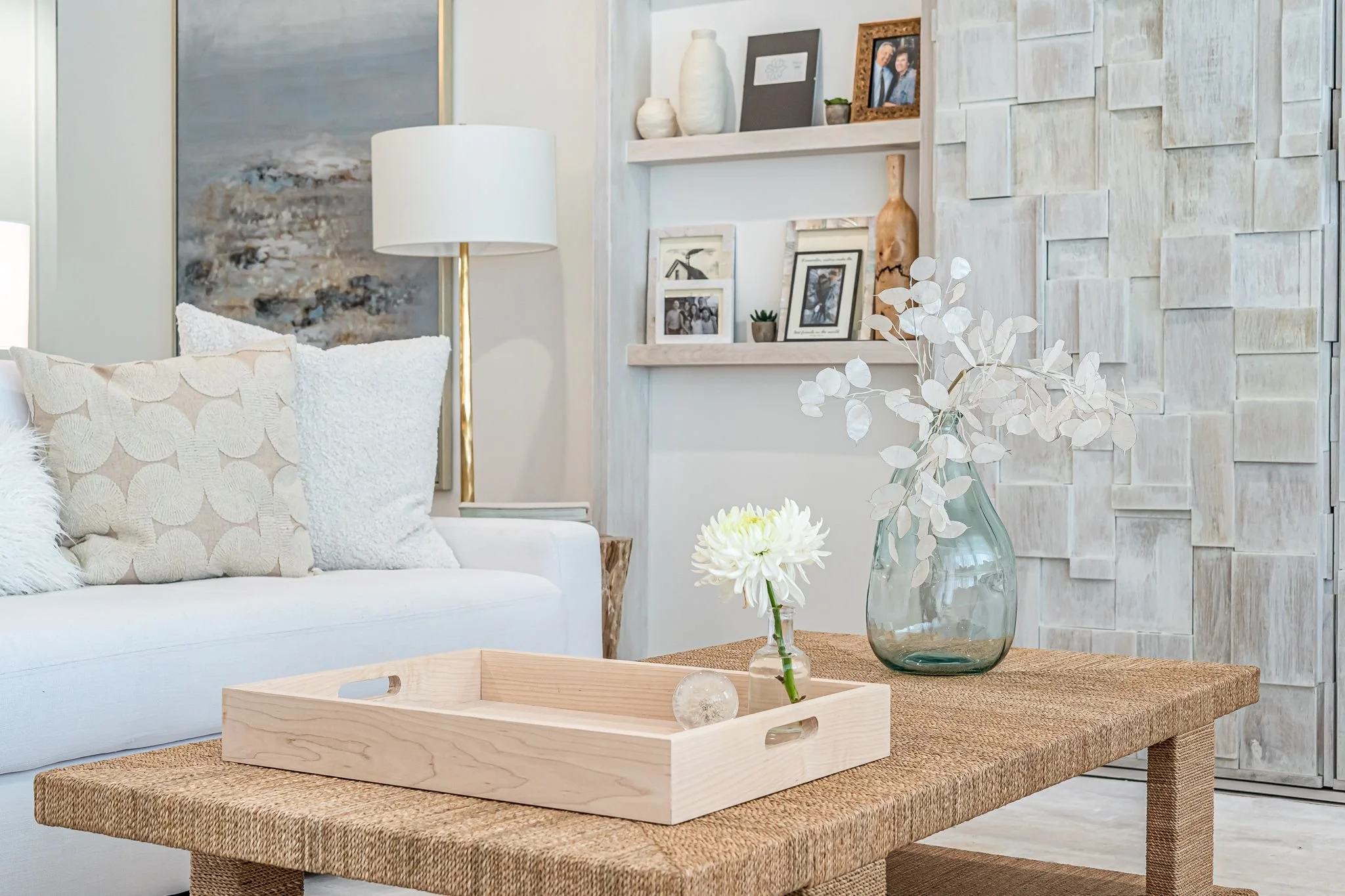 Living room with a white sofa decorated with beige, white, and fuzzy pillows, a wooden coffee table with a vase of white flowers and dried branches, a white shelf with framed photos and small decorative items, a tall lamp with a white shade and gold 