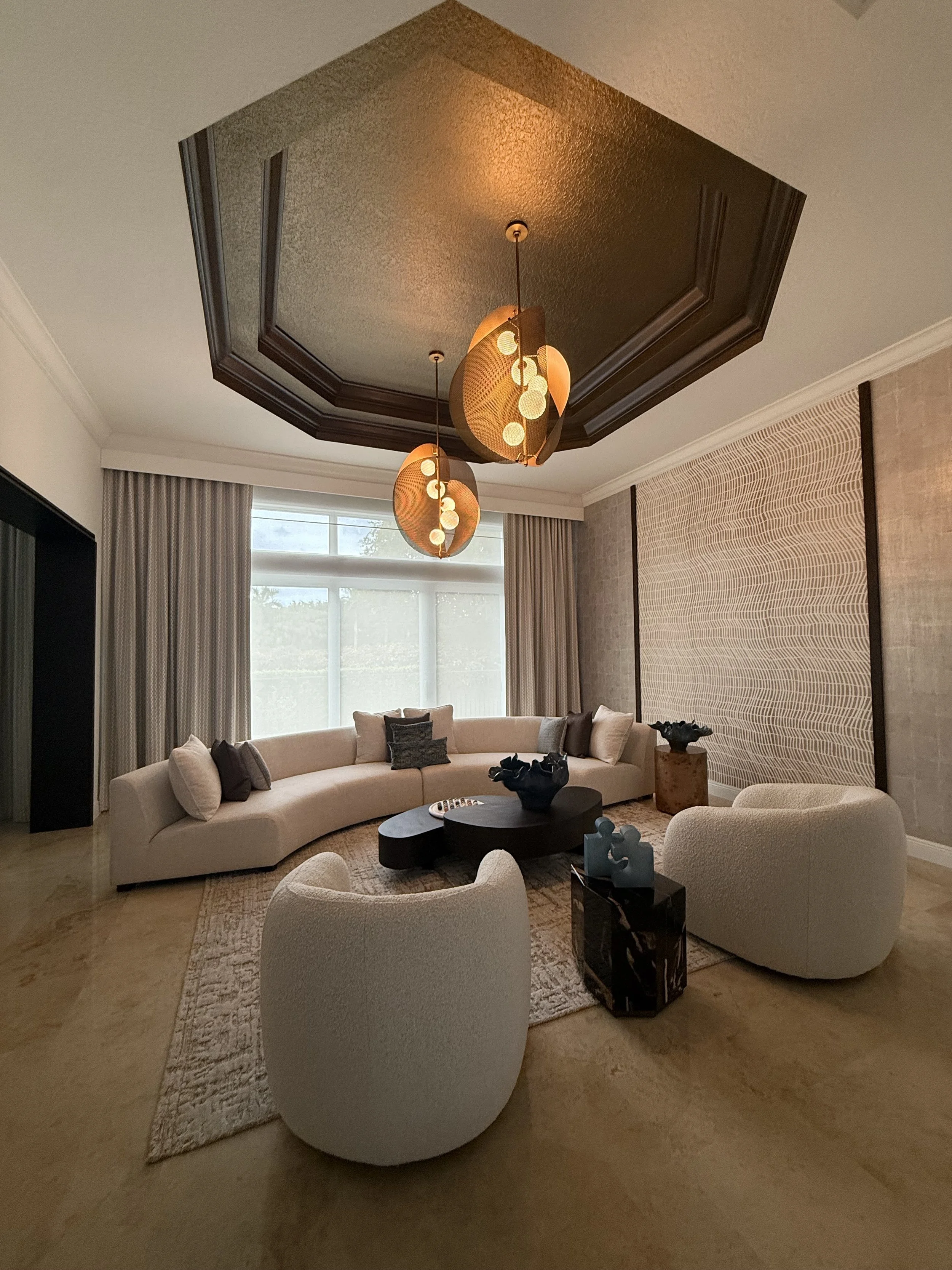 Living room with a curved white sectional sofa, two white rounded chairs, black coffee tables, decorative vases, large window with curtains, textured wall, and hanging pendant lights. Ceiling has a dark finish with decorative trim.